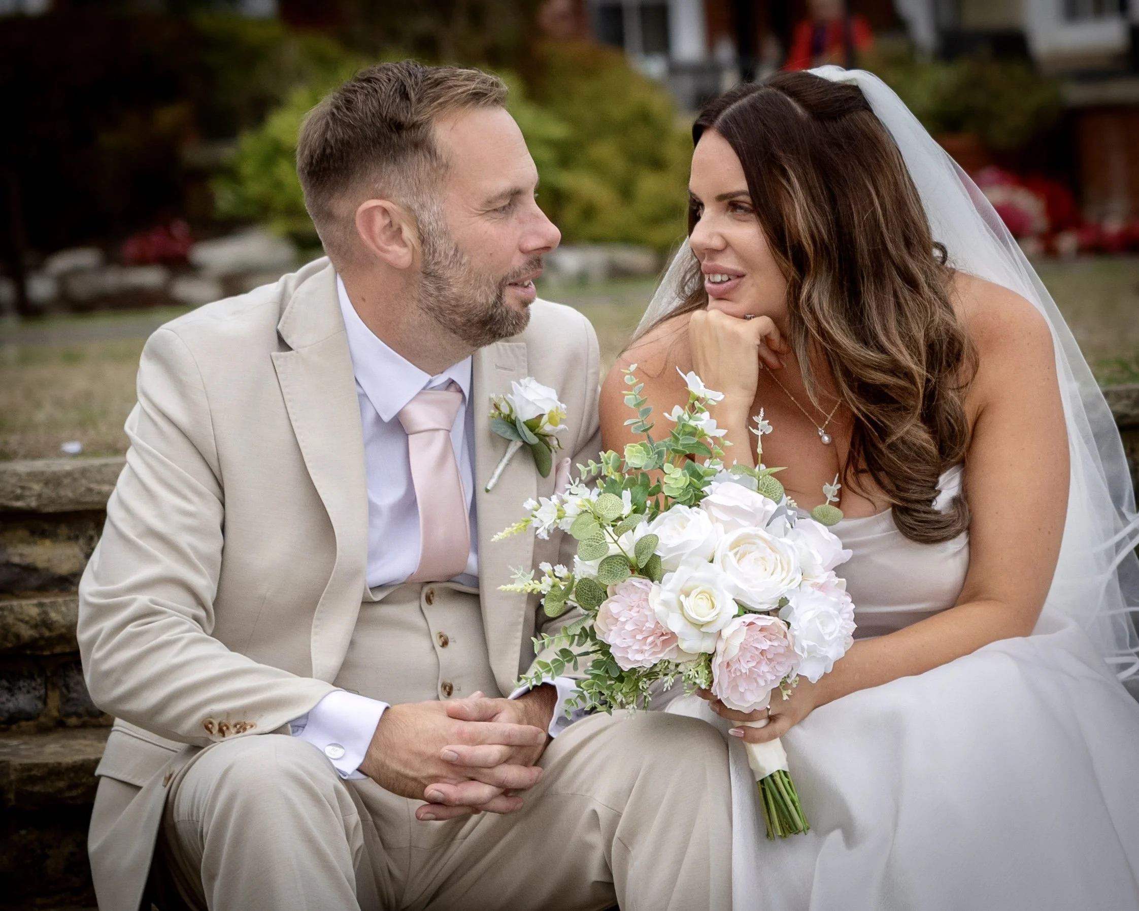 A bride and groom sitting on stairs outdoors during their wedding, facing each other, with the bride holding a bouquet of white and pink flowers. The groom is wearing a light-colored suit and the bride is wearing a white dress with a veil.