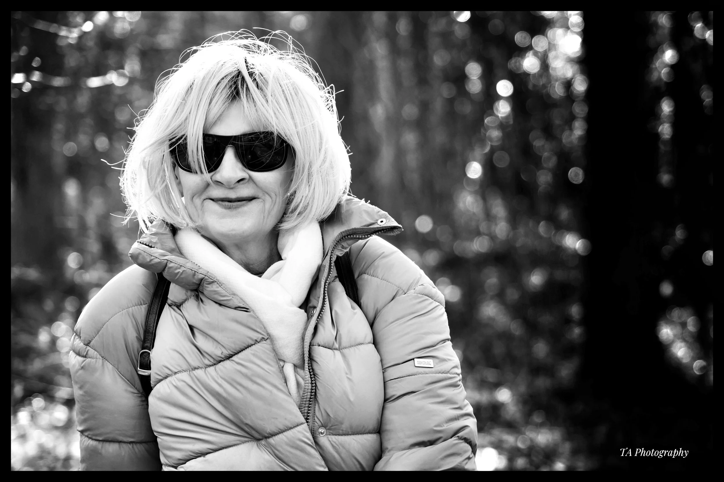 A black and white photo of a woman with blonde hair wearing sunglasses and a puffy jacket, standing outdoors with blurred trees and sunlight in the background.