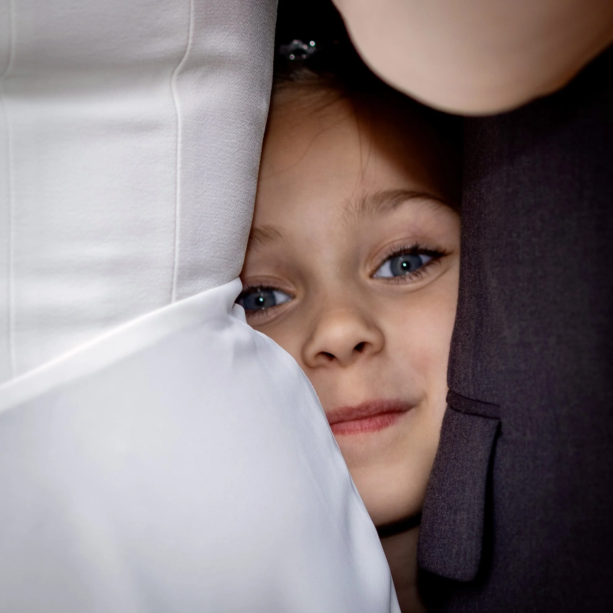 Close-up of a young person's face with blue eyes, partially covered by a shiny white fabric and a dark piece of clothing, with some strands of hair visible.