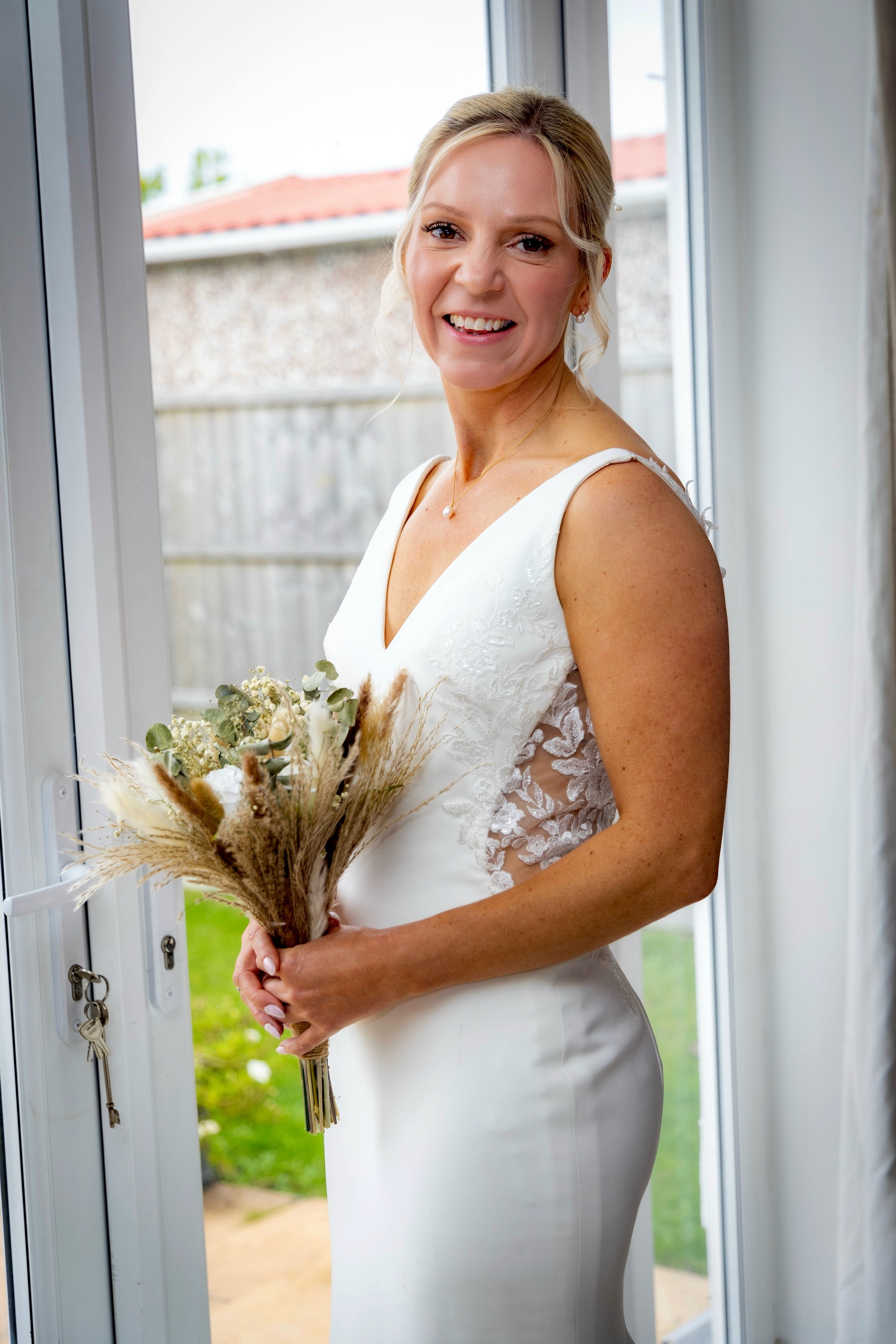 A smiling blonde woman in a white dress holding a bouquet of dried flowers stands near an open door, with a garden and house visible outside.