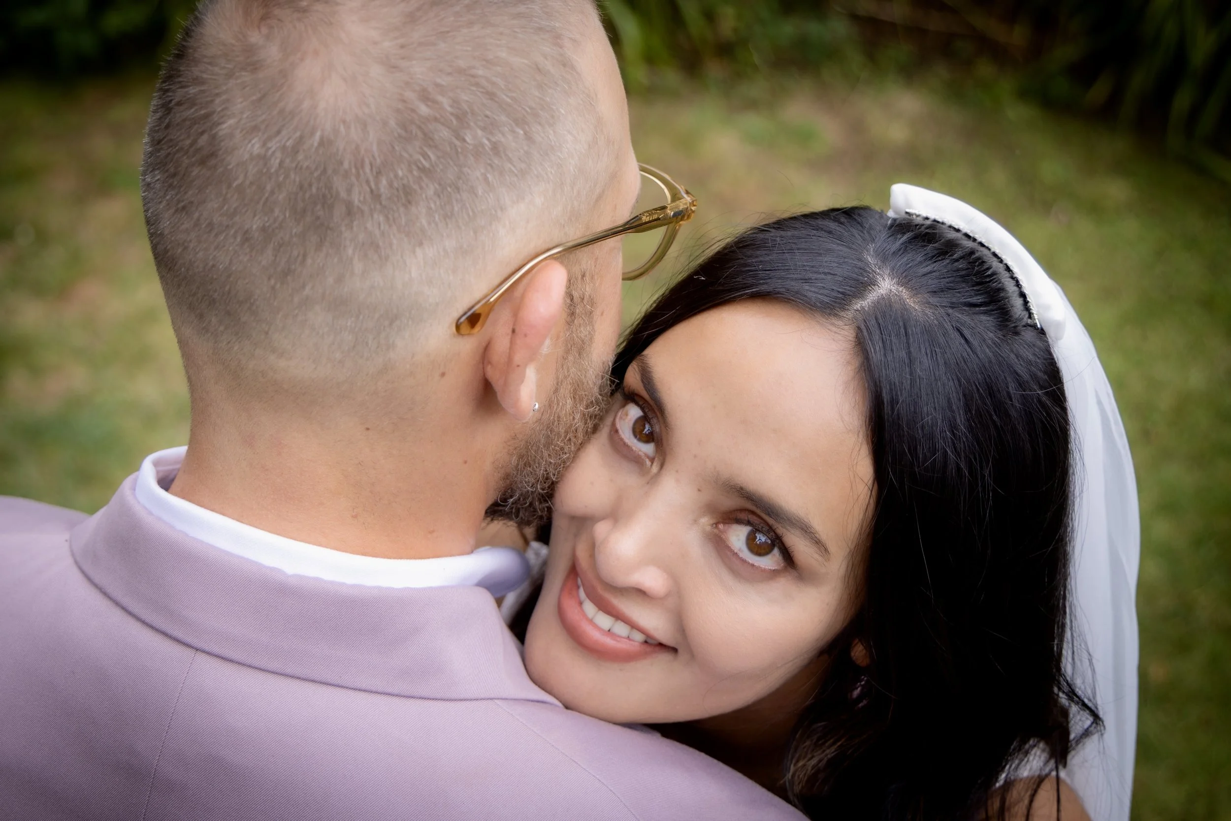 A couple embracing outdoors, with the woman looking up at the camera and smiling.