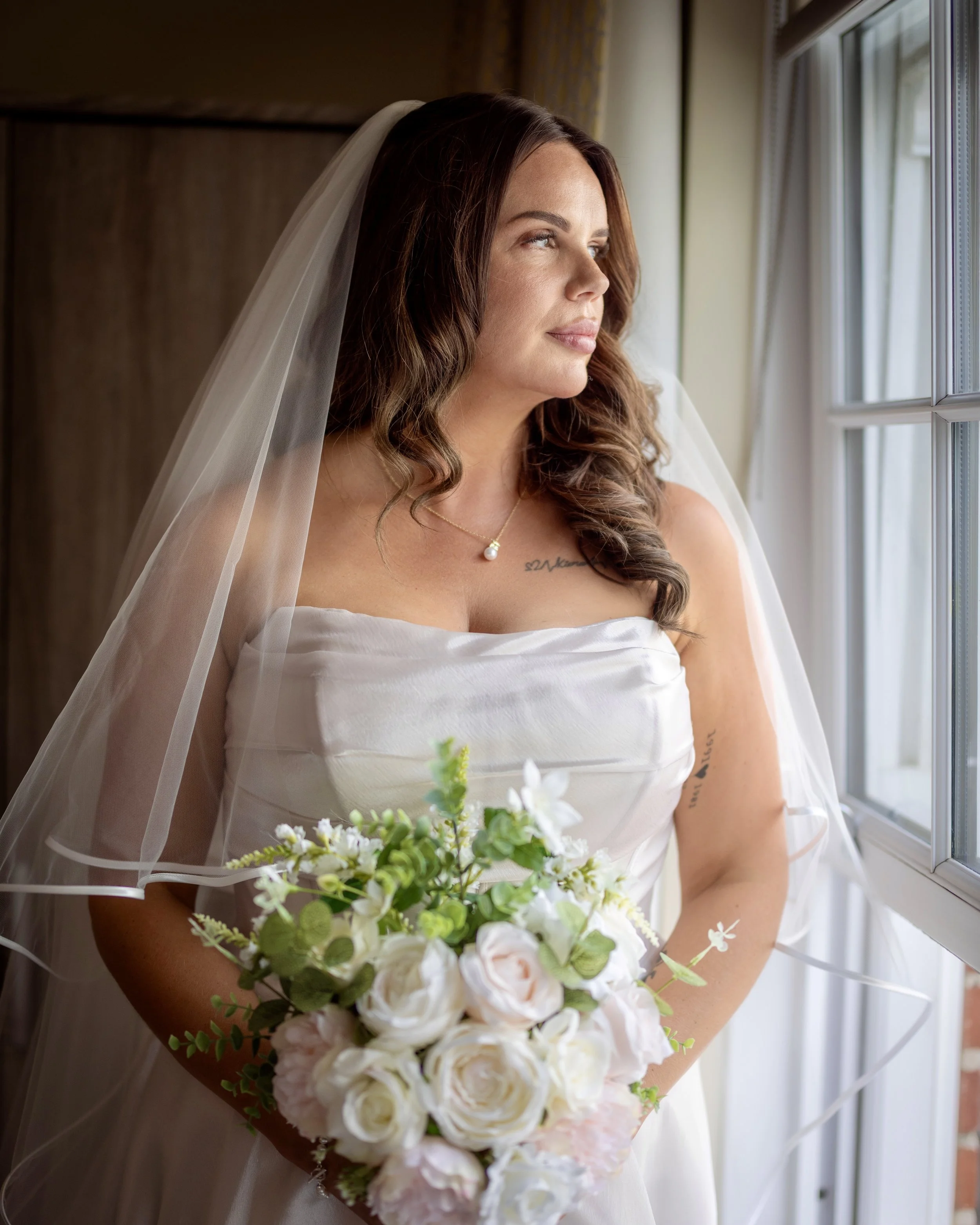 A woman in a wedding dress and veil holding a bouquet of white and pink flowers, looking out a window.