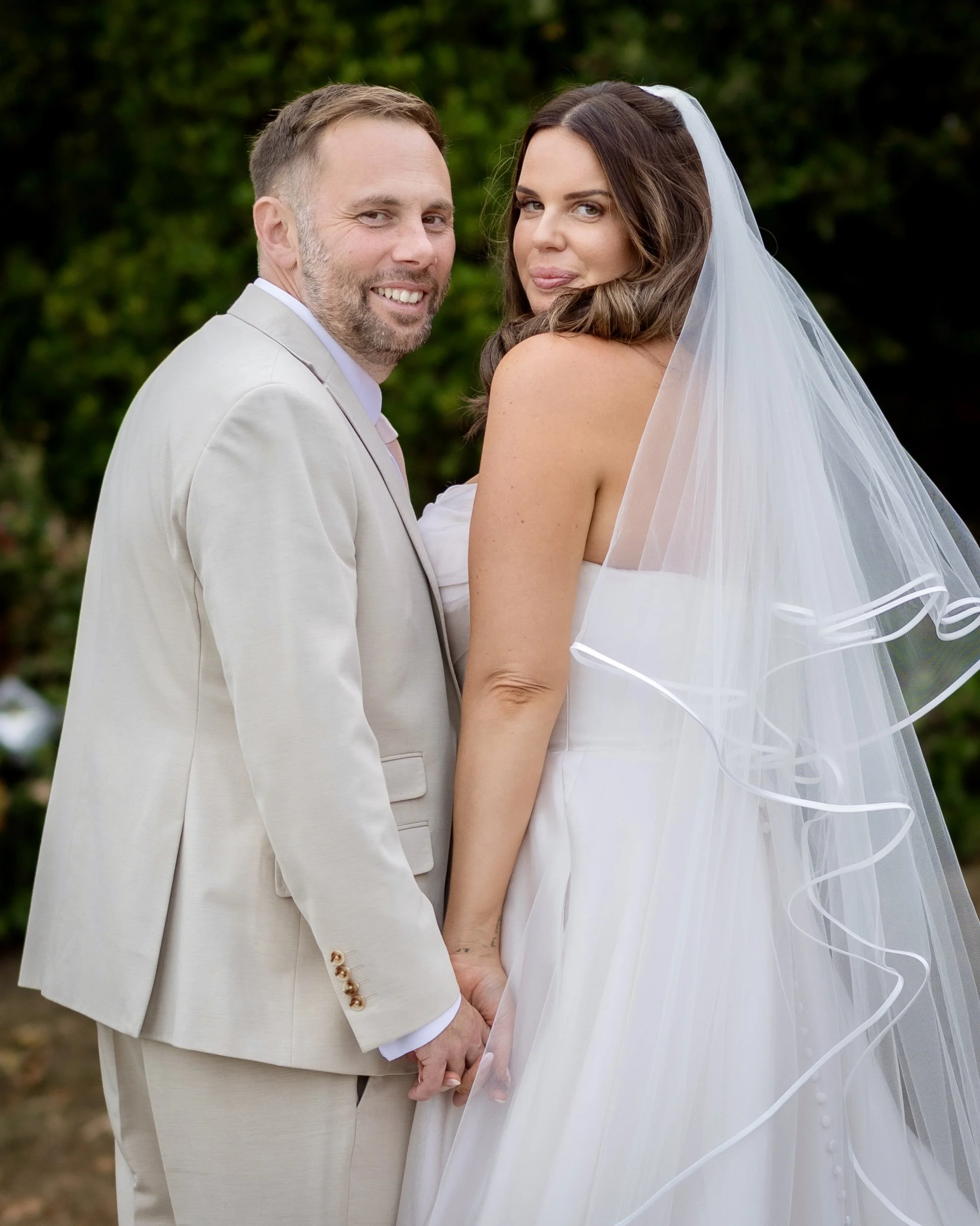 A newlywed couple holding hands outdoors, with the groom in a beige suit and the bride in a white wedding dress and veil, standing close and smiling at the camera.