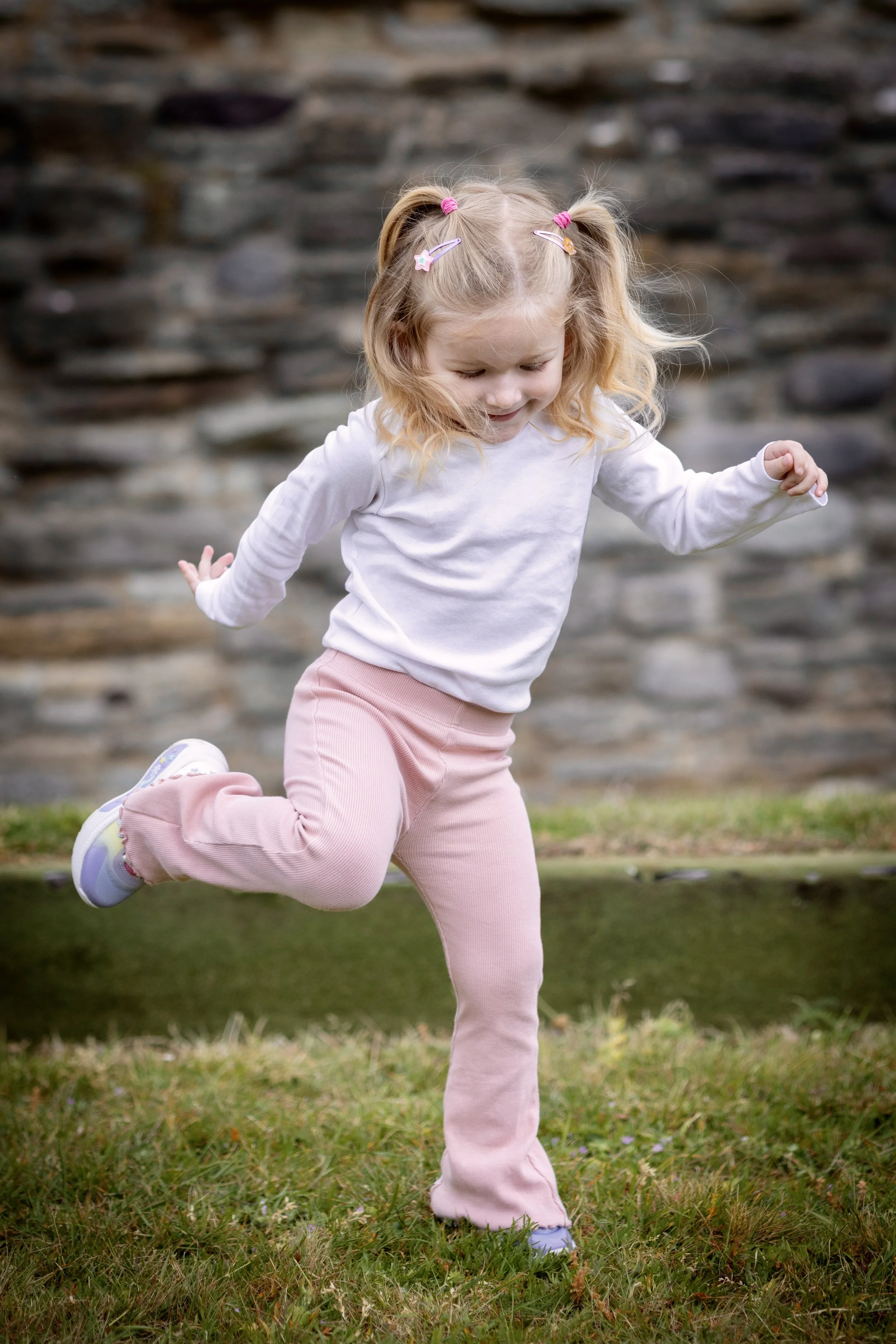 A young girl with blonde hair in pigtails, wearing hair clips, a white long-sleeve shirt, and pink pants, running on a grassy area in front of a stone wall.