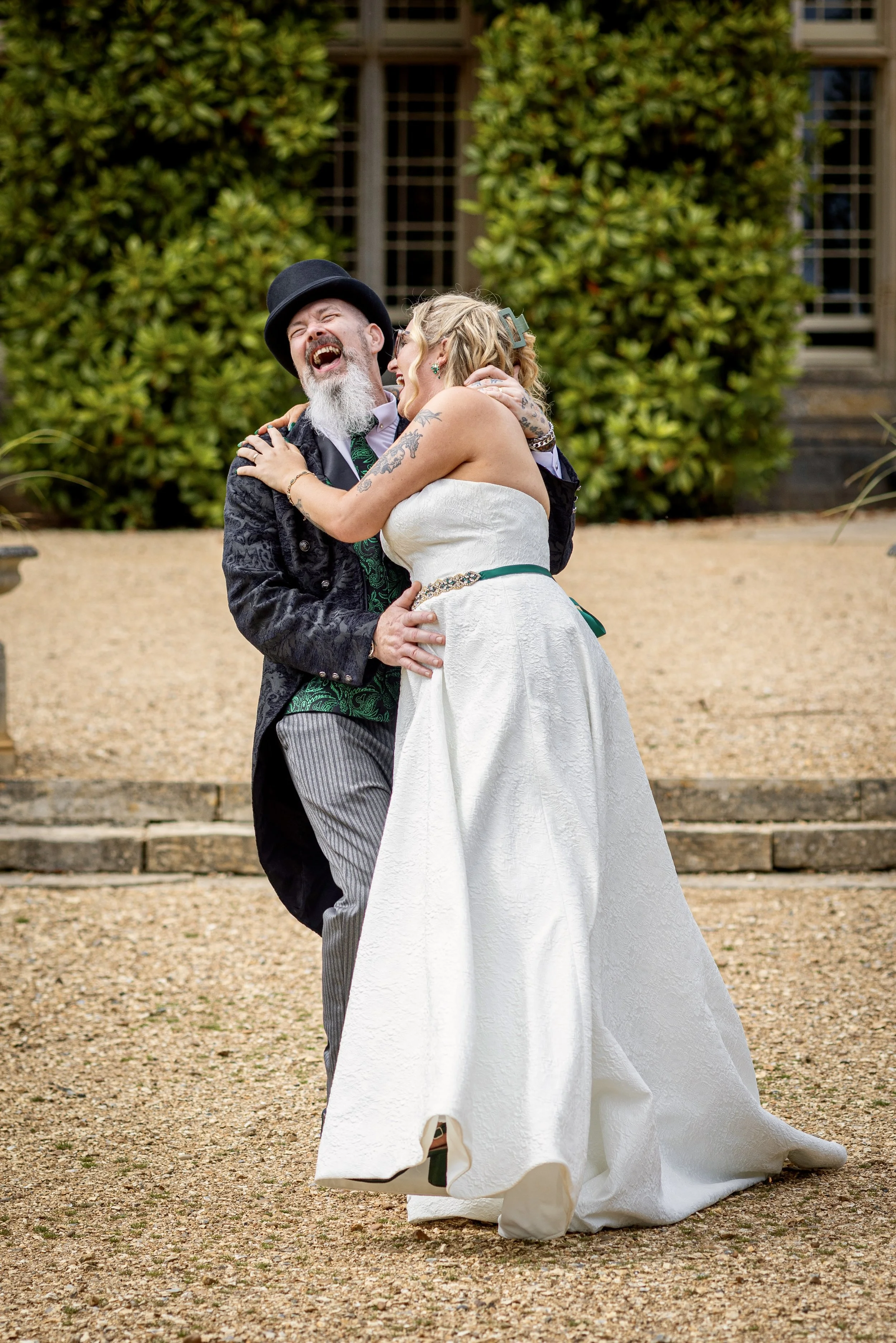 A couple dressed in wedding attire sharing a joyful dance outdoors, with a background of green shrubbery and a historic-looking building.