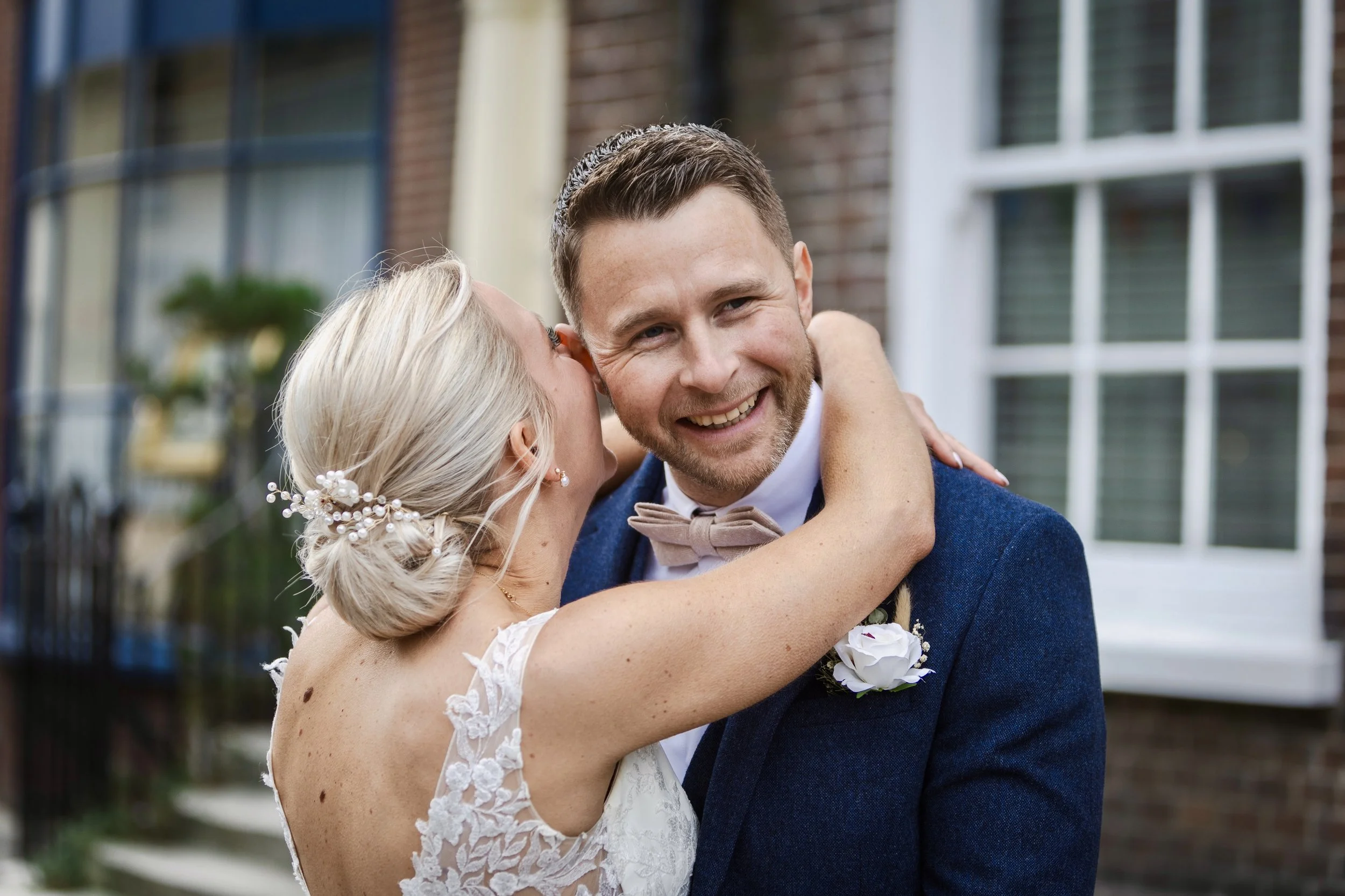 A bride and groom sharing a joyful hug outside a building with brick walls and large windows.