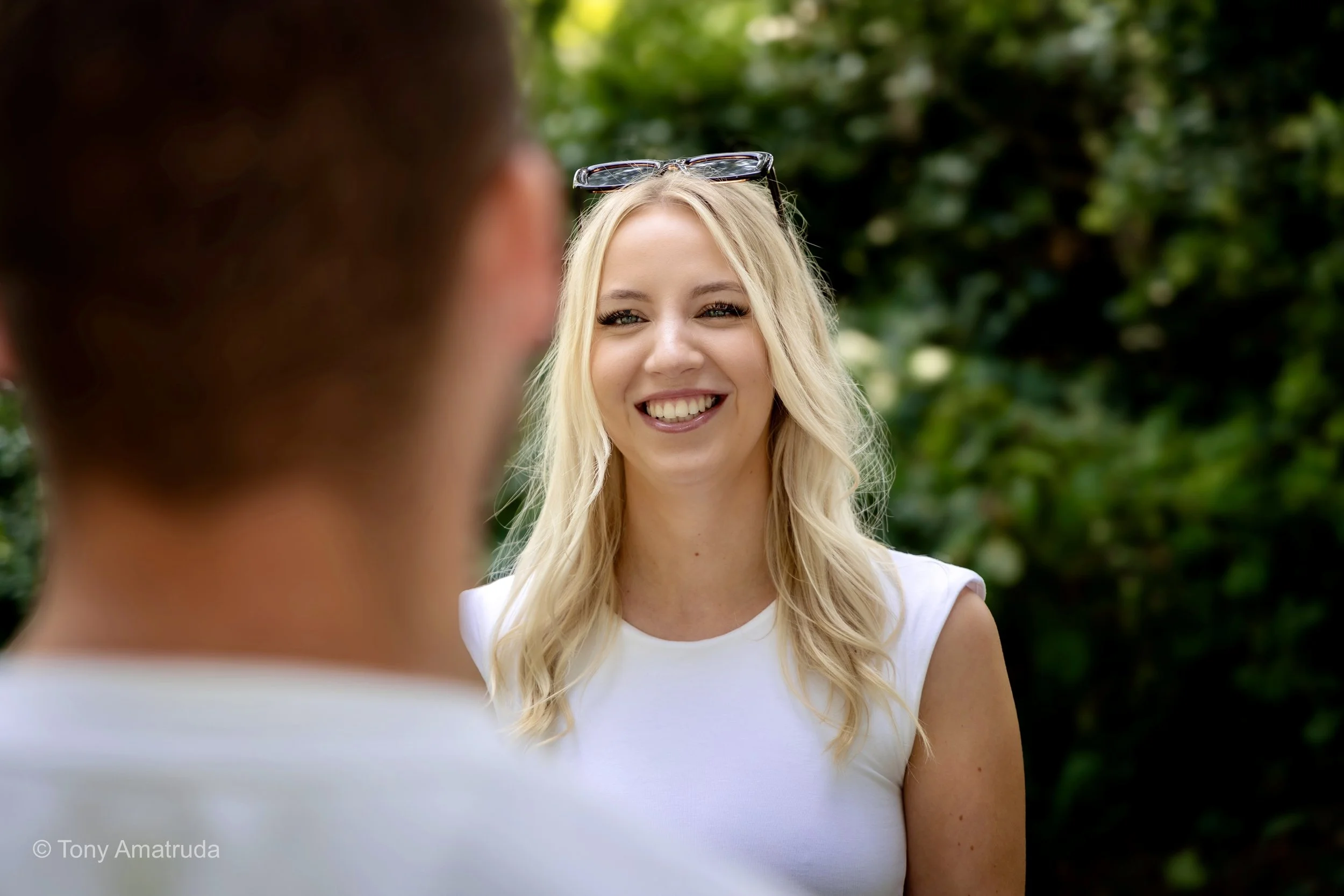 A young woman with long blonde hair smiling, wearing a white tank top, sunglasses on her head, outdoors with green foliage in the background.