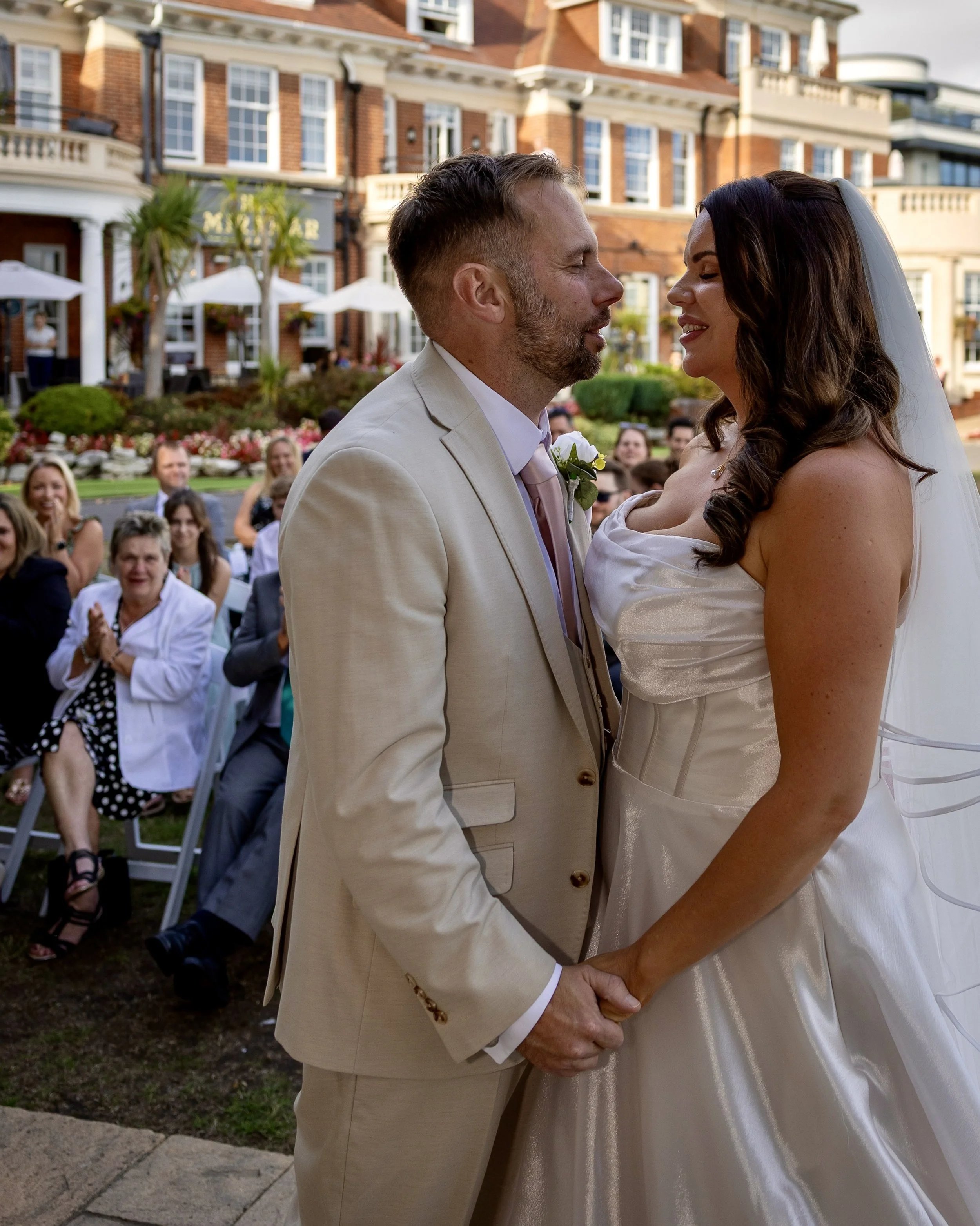 A couple is standing close together, holding hands, during their wedding ceremony outdoors. The groom has short light brown hair and a beard, wearing a cream-colored suit with a white shirt and a pink tie. The bride has long dark brown hair, wearing 