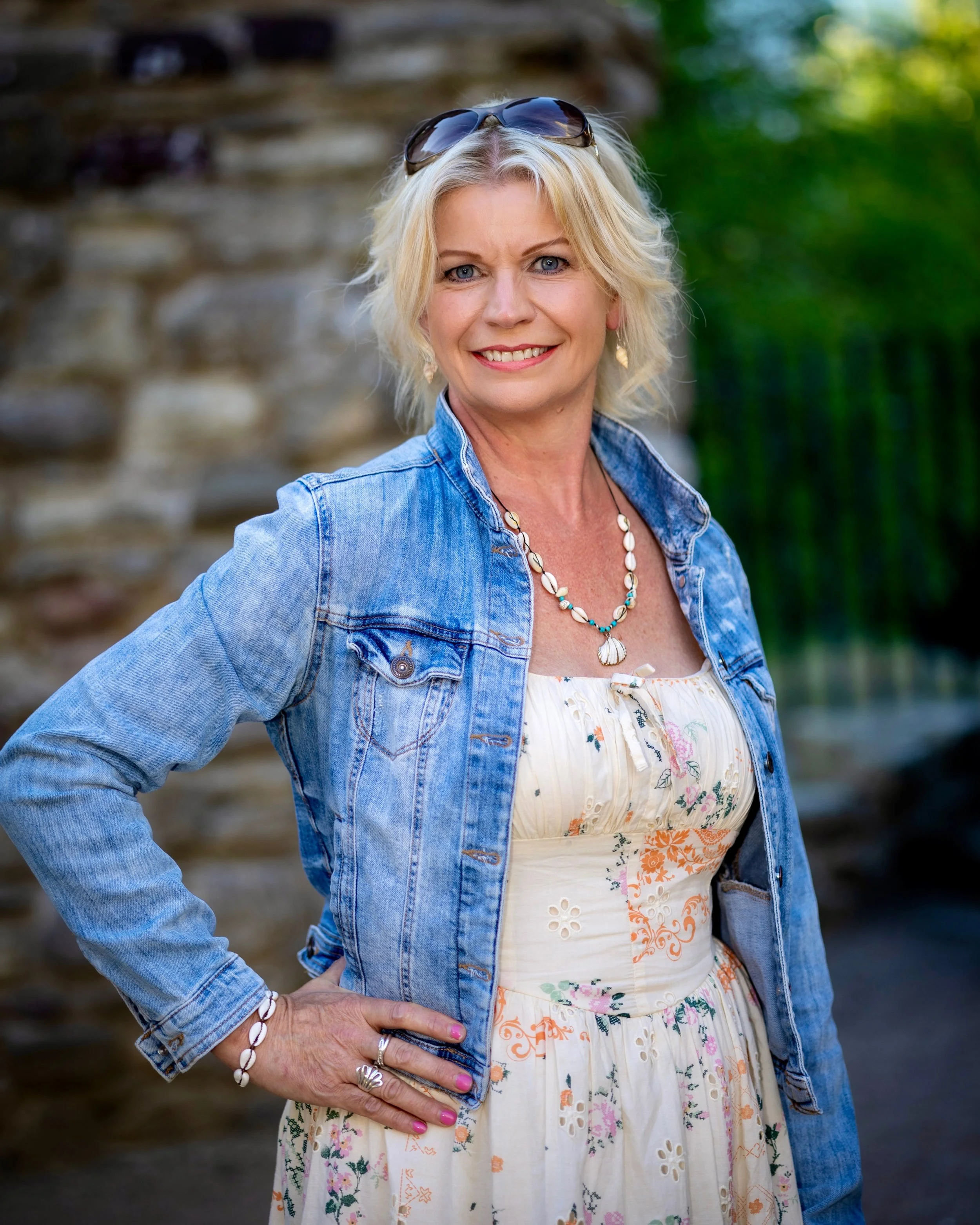 A middle-aged woman with blonde hair, wearing sunglasses on her head, a denim jacket, and a white floral dress. She is standing outdoors with her hand on her hip, smiling at the camera. The background features blurred natural scenery with trees and r
