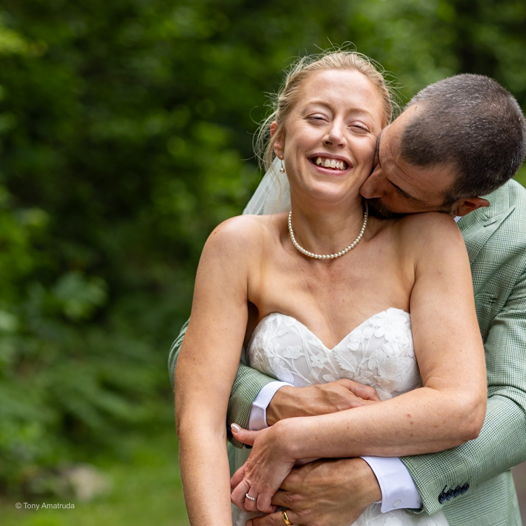 A woman smiling and laughing while hugging a man outdoors surrounded by greenery.