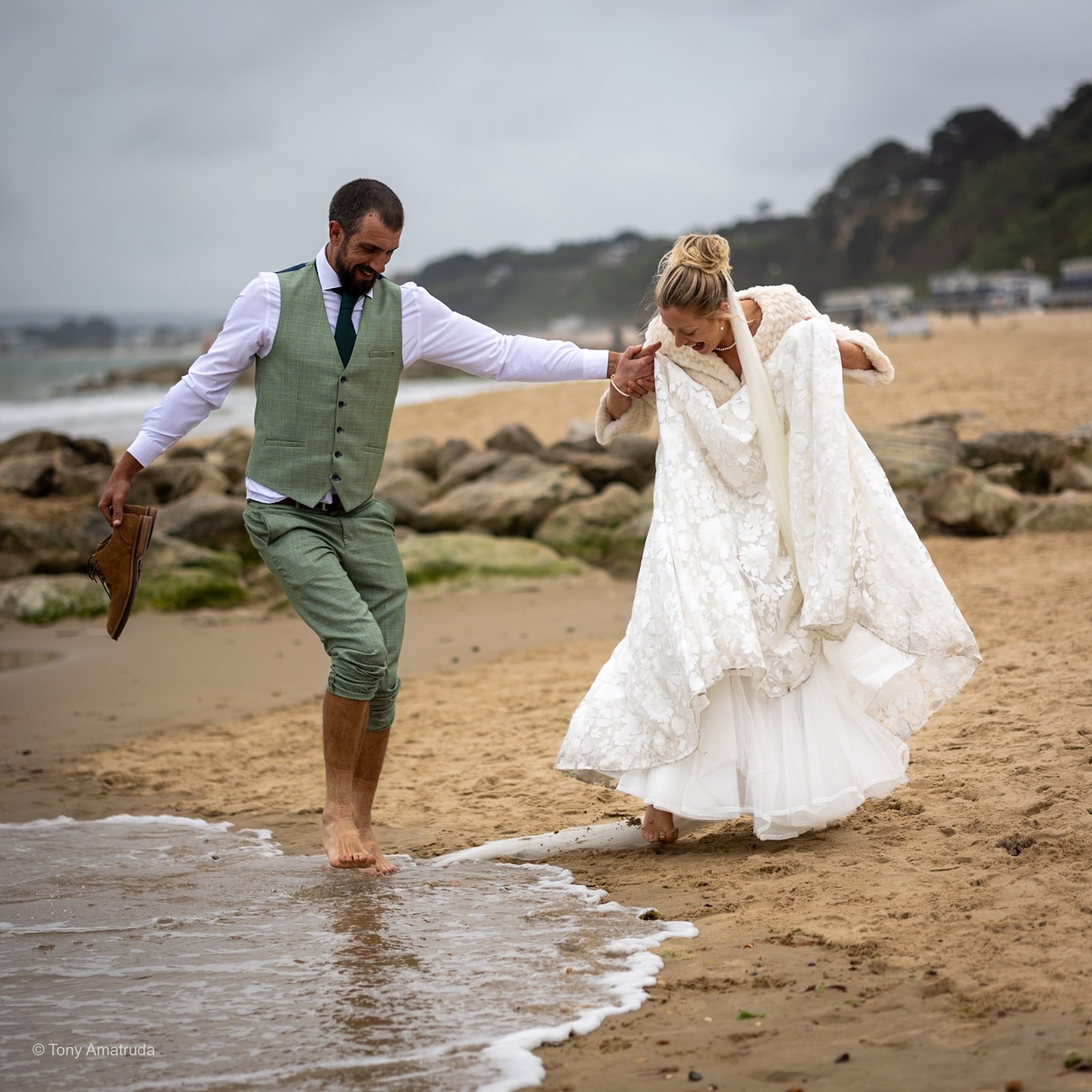 A man and woman dressed in wedding attire walking barefoot along a beach, holding hands and laughing.