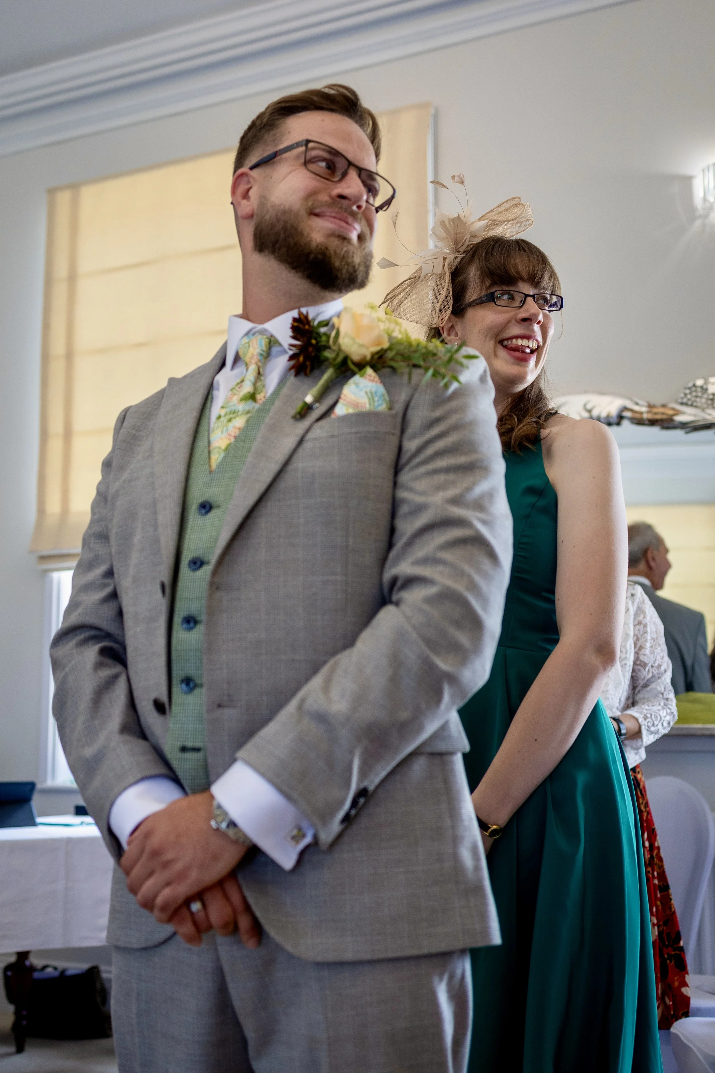 Man in a light gray suit with flower boutonniere standing next to woman in a teal dress with fascinator at a wedding or formal event.