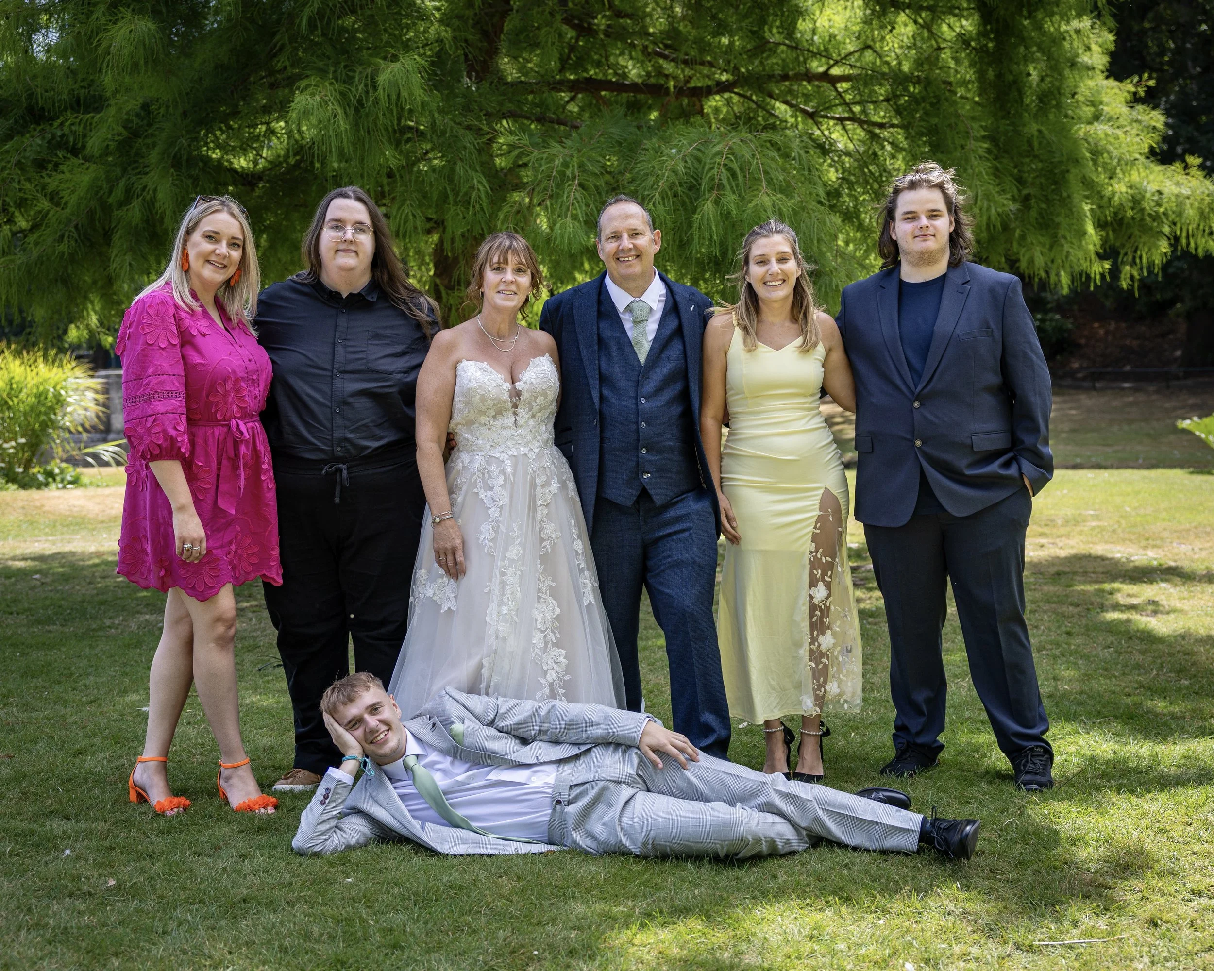 Group of six adults and one child posing outdoors on a grassy area with green trees in the background, smiling and dressed in formal and semi-formal attire.