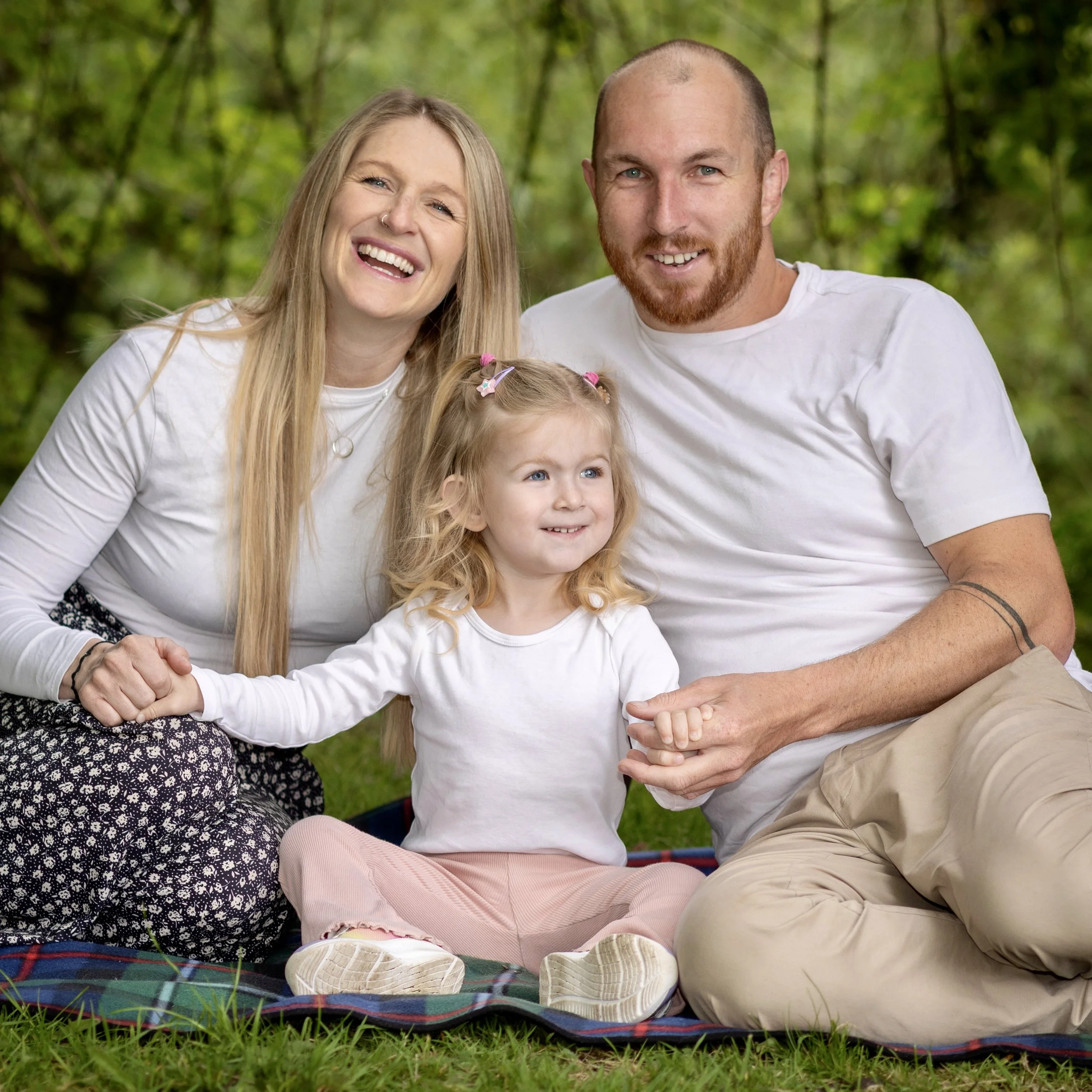 A family of three sitting on a plaid blanket outdoors, surrounded by green grass and trees, smiling and looking happy.
