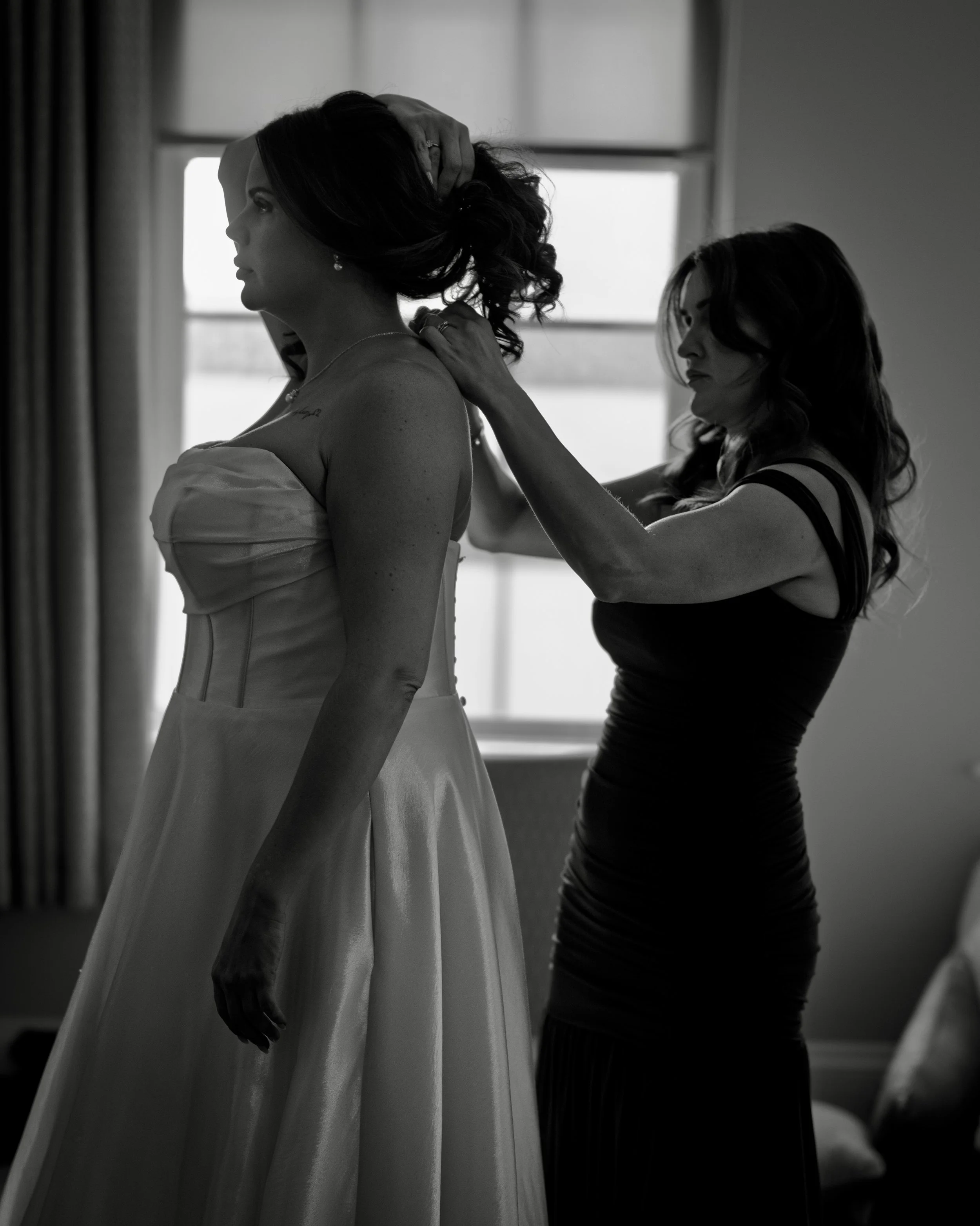 Black and white photo of a woman in a wedding dress having her hair styled by another woman in a sleeveless dress, with a window in the background.