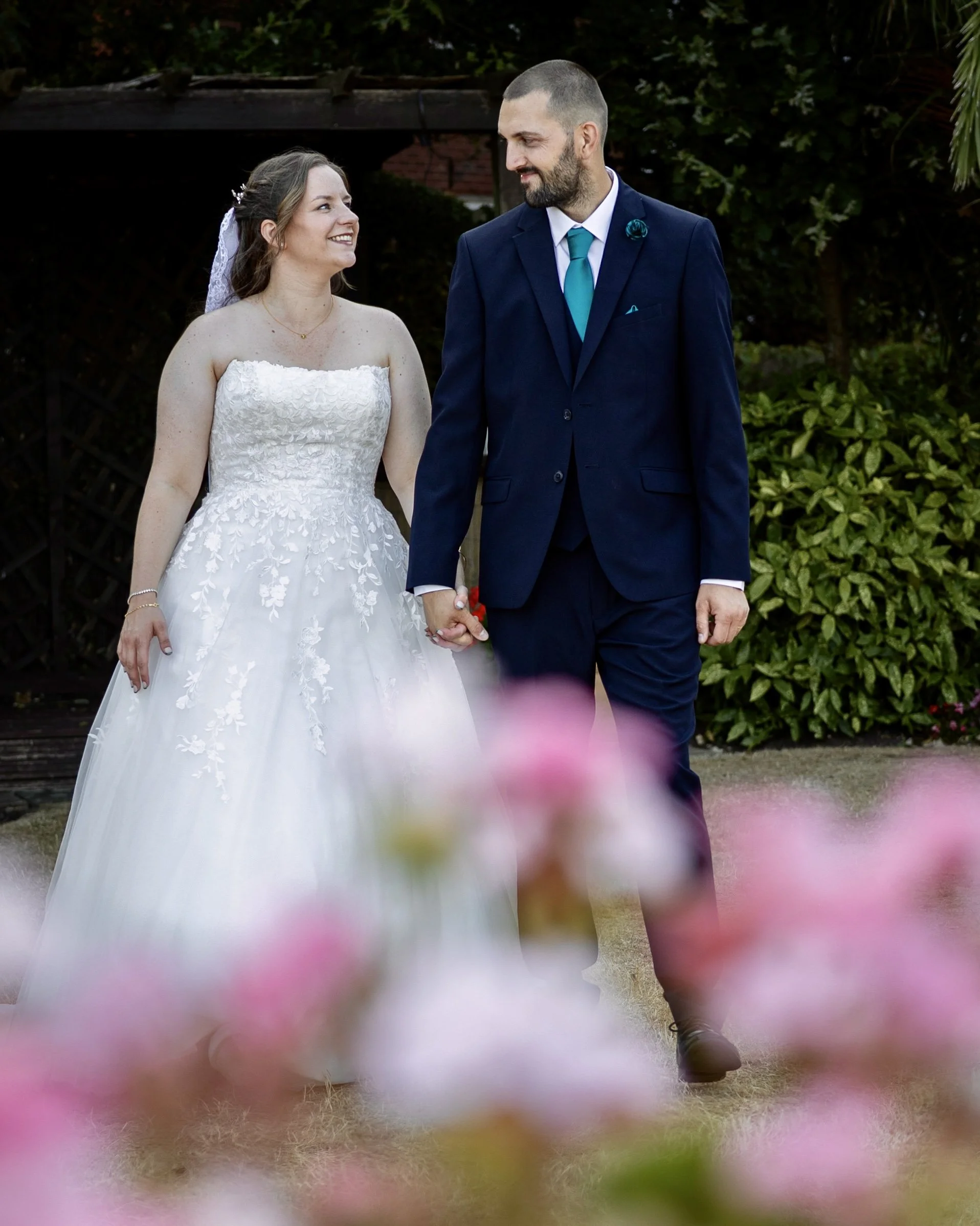 A bride and groom holding hands and smiling at each other outdoors, with a background of greenery and a wooden structure.