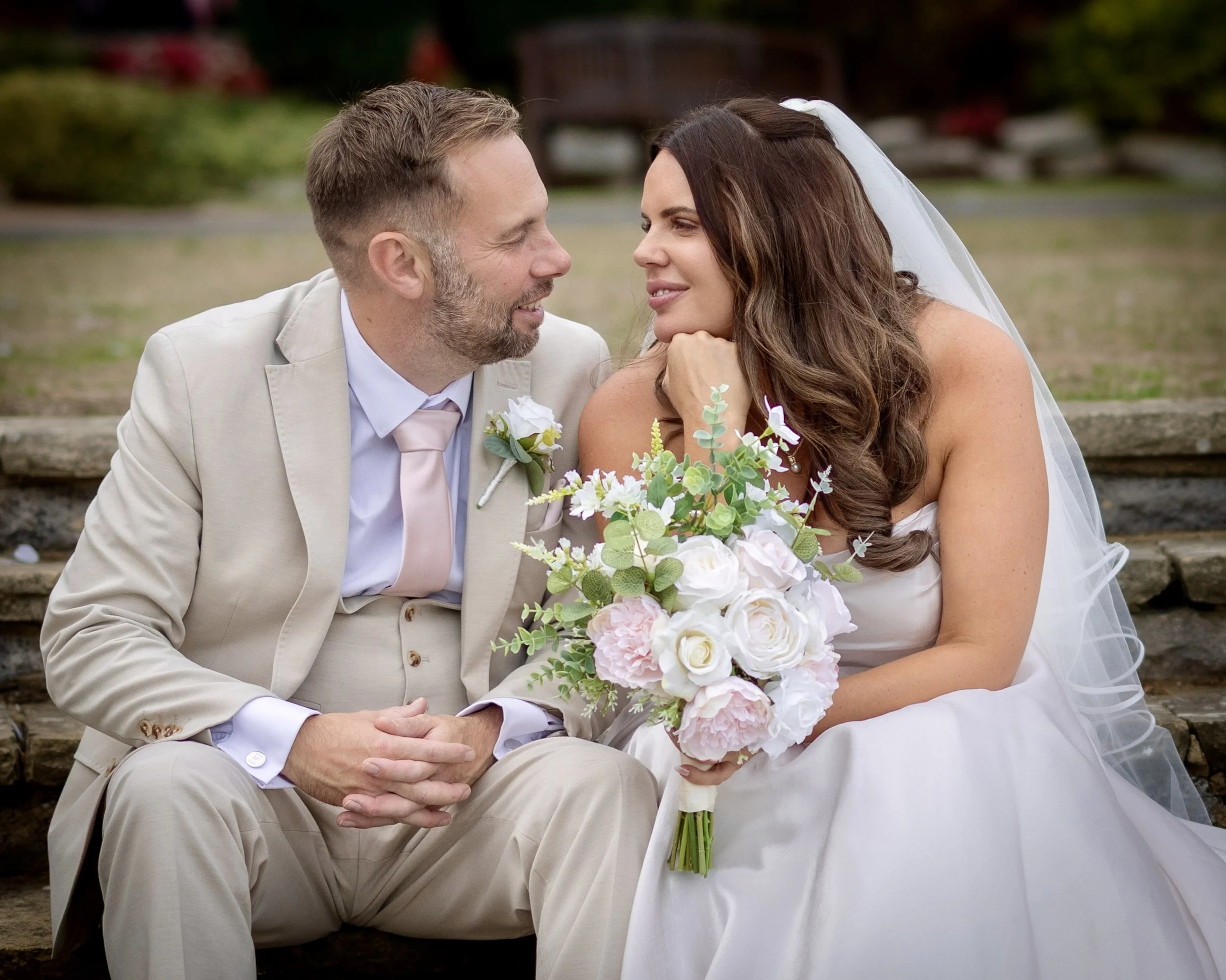 A bride and groom sitting outdoors. The bride, with long brown hair and wearing a veil, holds a bouquet of flowers. The groom, with short hair and a beard, dressed in a beige suit, sits close, looking at each other with smiles.