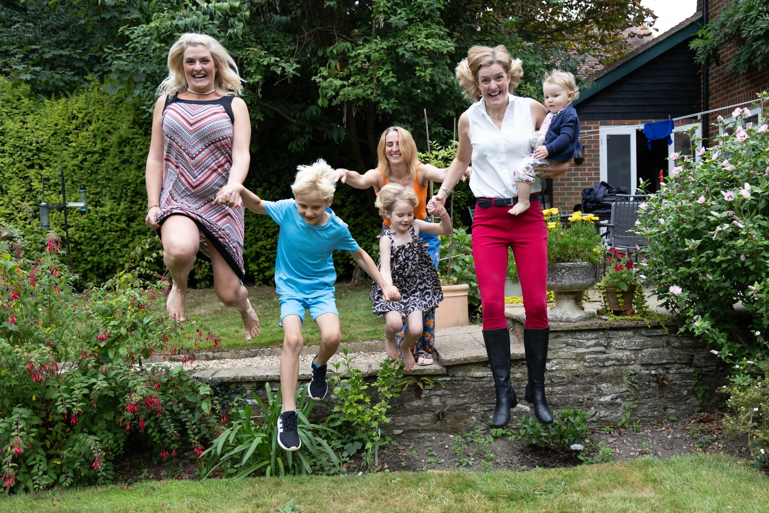 Group of smiling women and children playing and jumping in a garden with plants, flowers, and a brick house in the background.