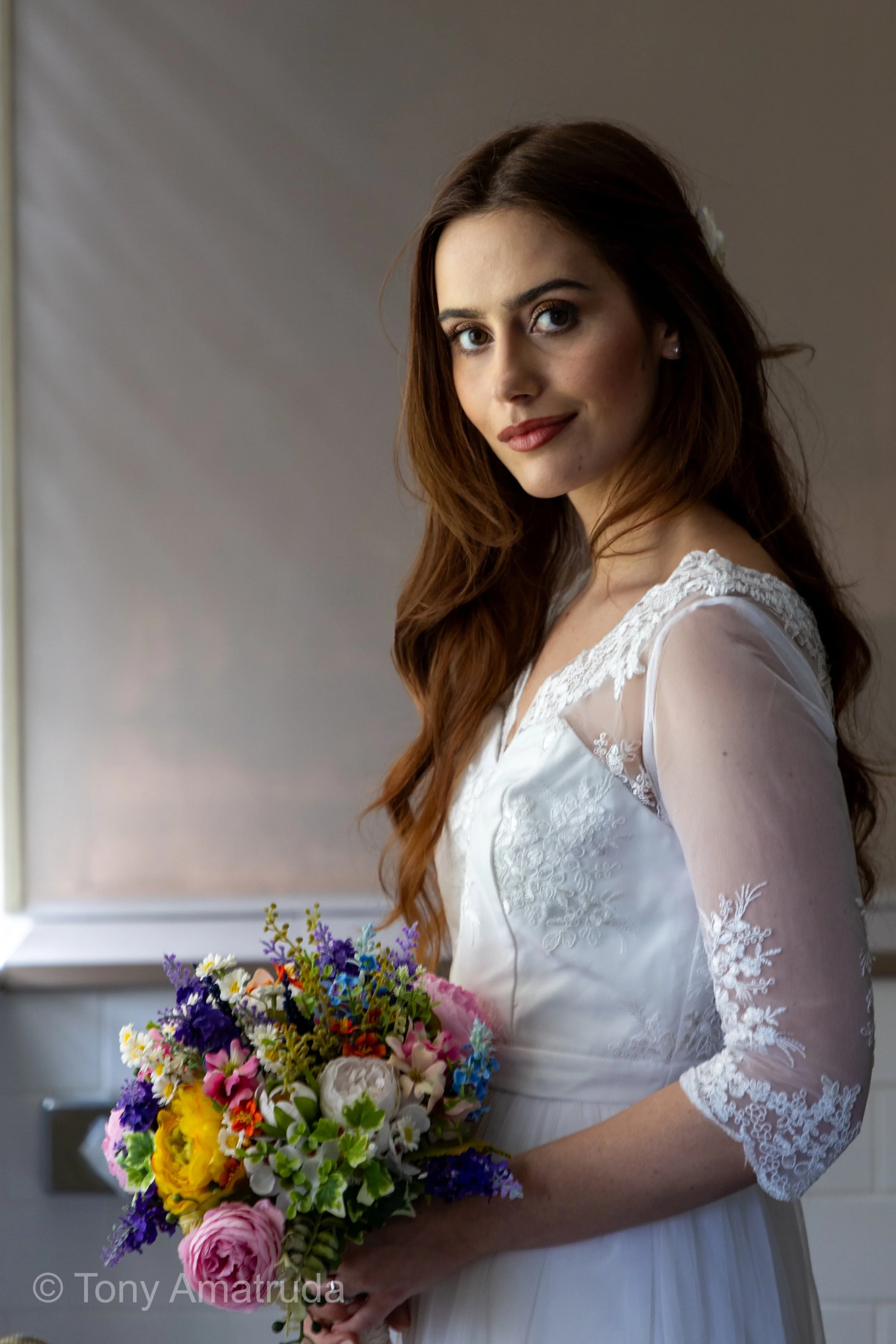 A woman with long, wavy brown hair wearing a white lace wedding dress, holding a colorful bouquet of flowers, standing indoors near a window.