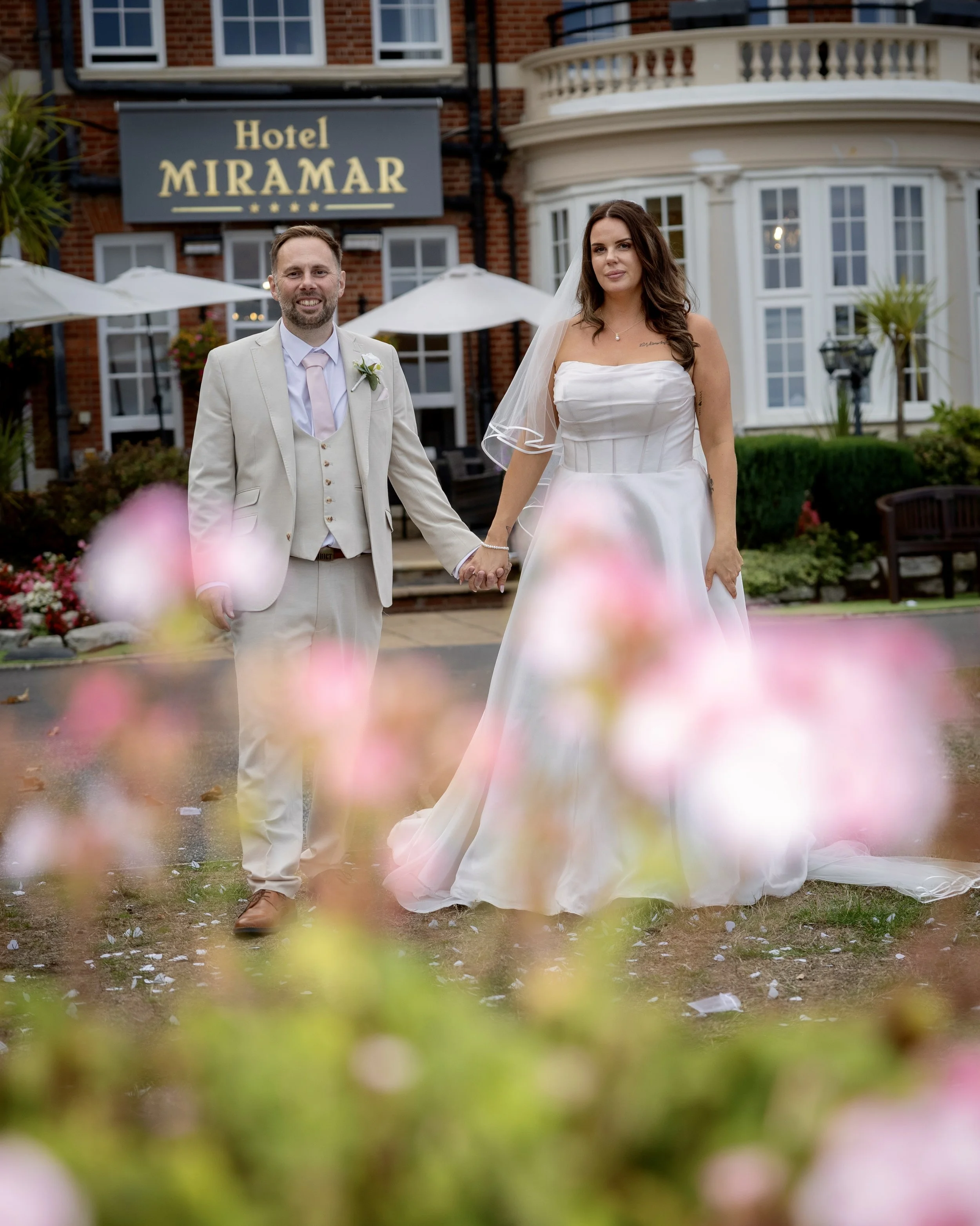 A newlywed couple holding hands in front of the Hotel Miramar during daytime. The groom wears a light suit with a boutonnière, and the bride wears a strapless white wedding dress with a veil. There are pink flowers and greenery in the foreground, wit