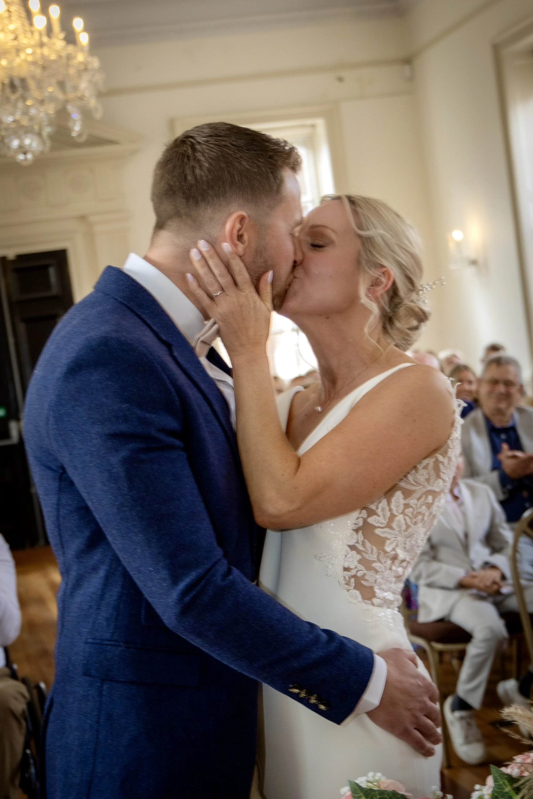 A bride and groom share a kiss during their wedding ceremony in a decorated indoor space with chandeliers, guests sitting in the background, and sunlight coming through windows.