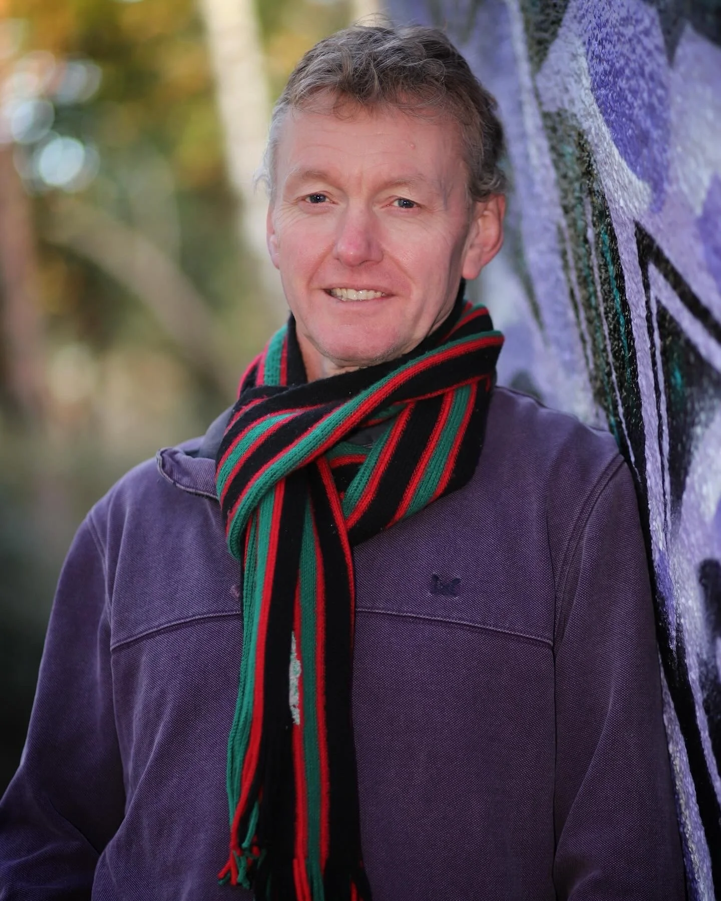 A man with light brown hair, wearing a purple jacket and a black scarf with red and green stripes, stands outdoors next to a graffiti-covered wall with blurred trees in the background.