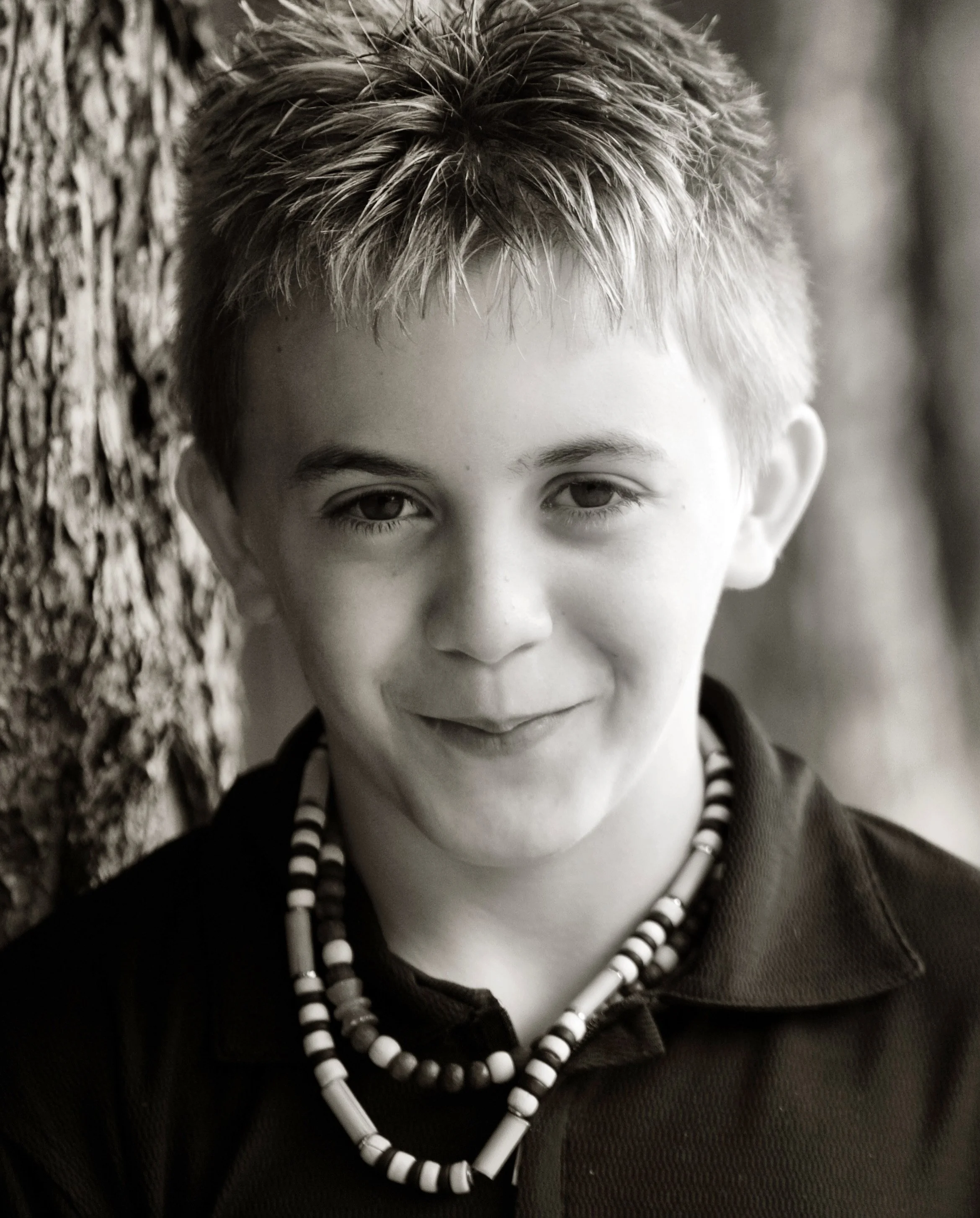 Close-up grayscale photograph of a young person with short, spiky hair, wearing a beaded necklace, standing next to a textured tree bark surface.