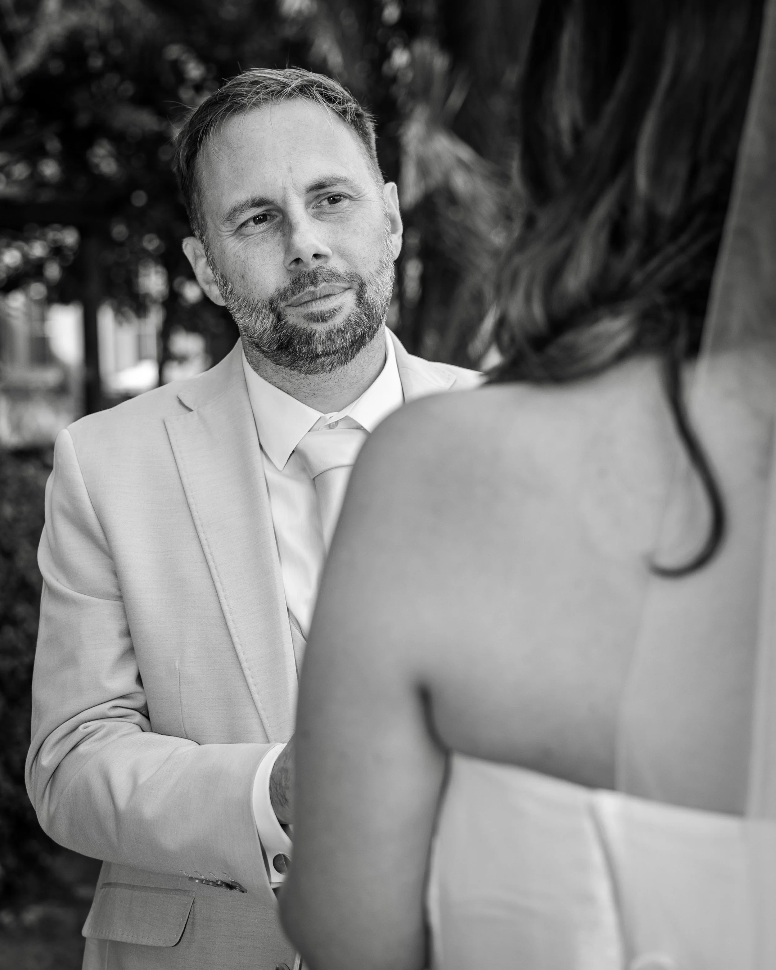 A man with a beard in a light-colored suit looking at a woman with long hair, seen from behind, outdoors.