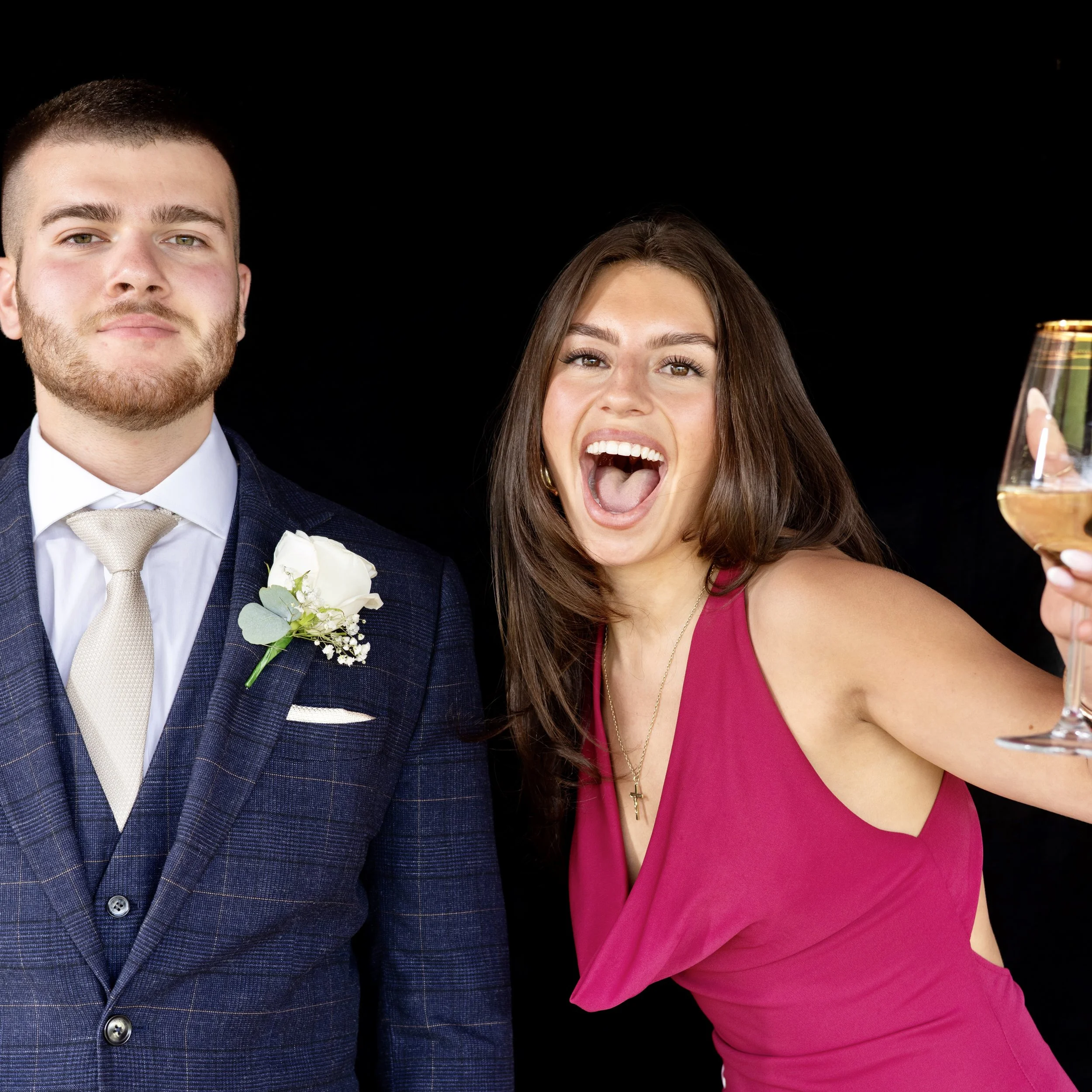 A man in a blue suit with a white shirt and beige tie standing next to a woman in a hot pink dress, taking a selfie with a smartphone in front of a black background.