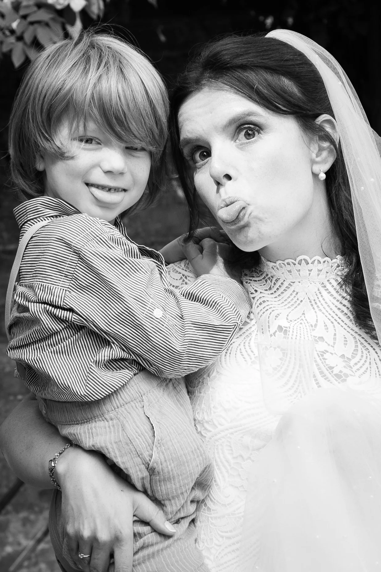 A woman in wedding attire making a funny face with her tongue out, holding a young boy with shaggy hair, smiling and sticking out his tongue. They are outdoors.