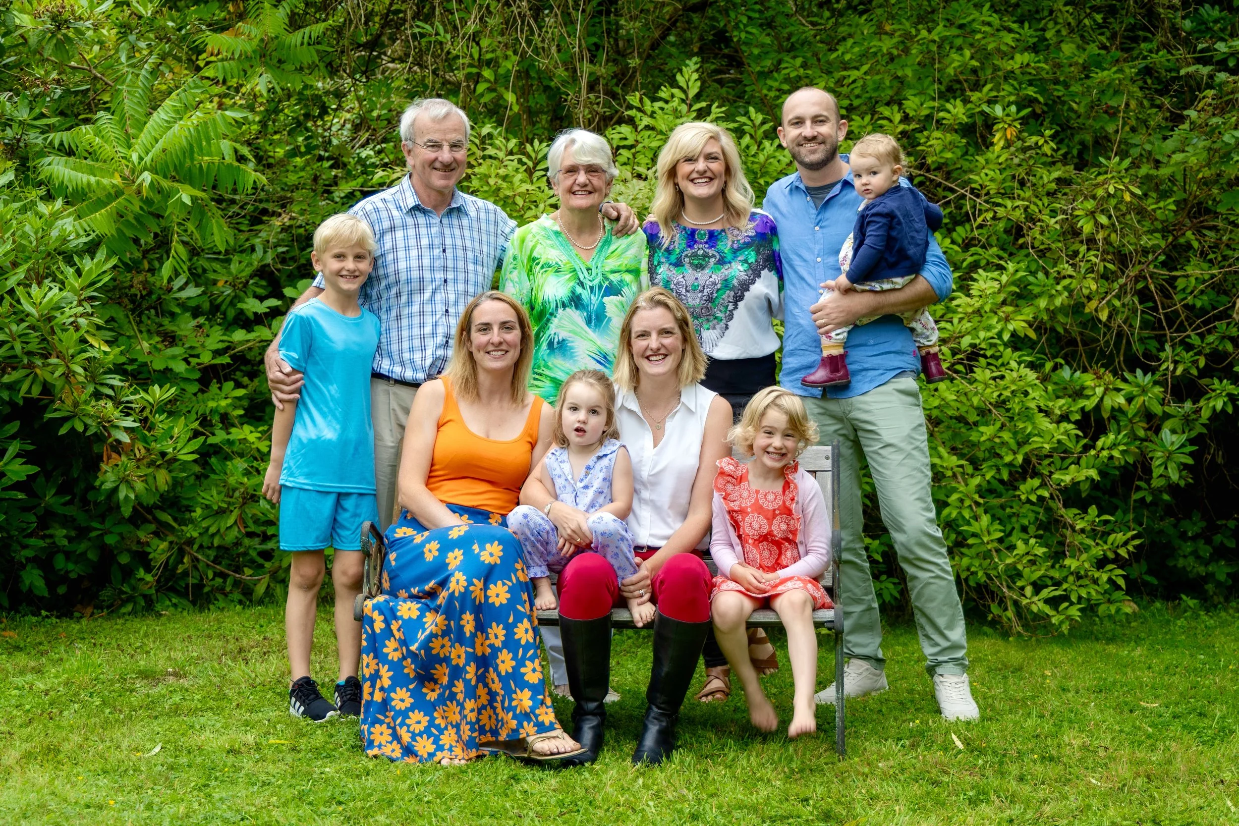 Family and friends gathered outdoors in a lush, green garden, smiling at the camera with some seated and some standing. There are children, adults, and elderly individuals, all dressed casually.