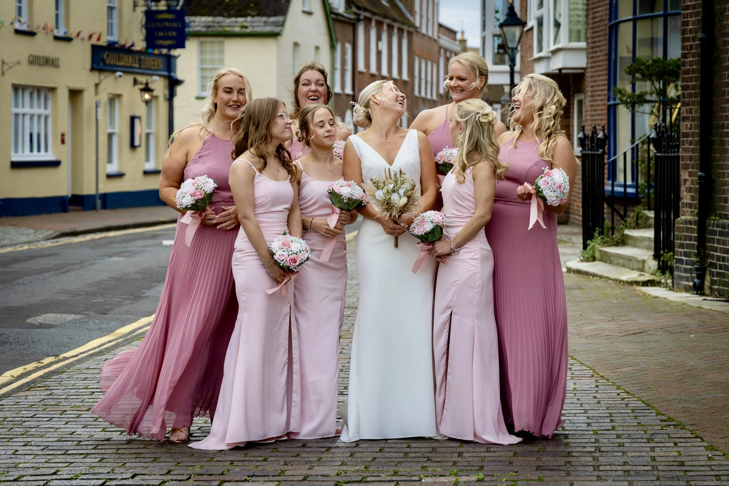A bride and seven bridesmaids standing on a street, all smiling and laughing. The bridesmaids wear pink dresses, and the bride wears a white dress. They each hold bouquets of flowers.