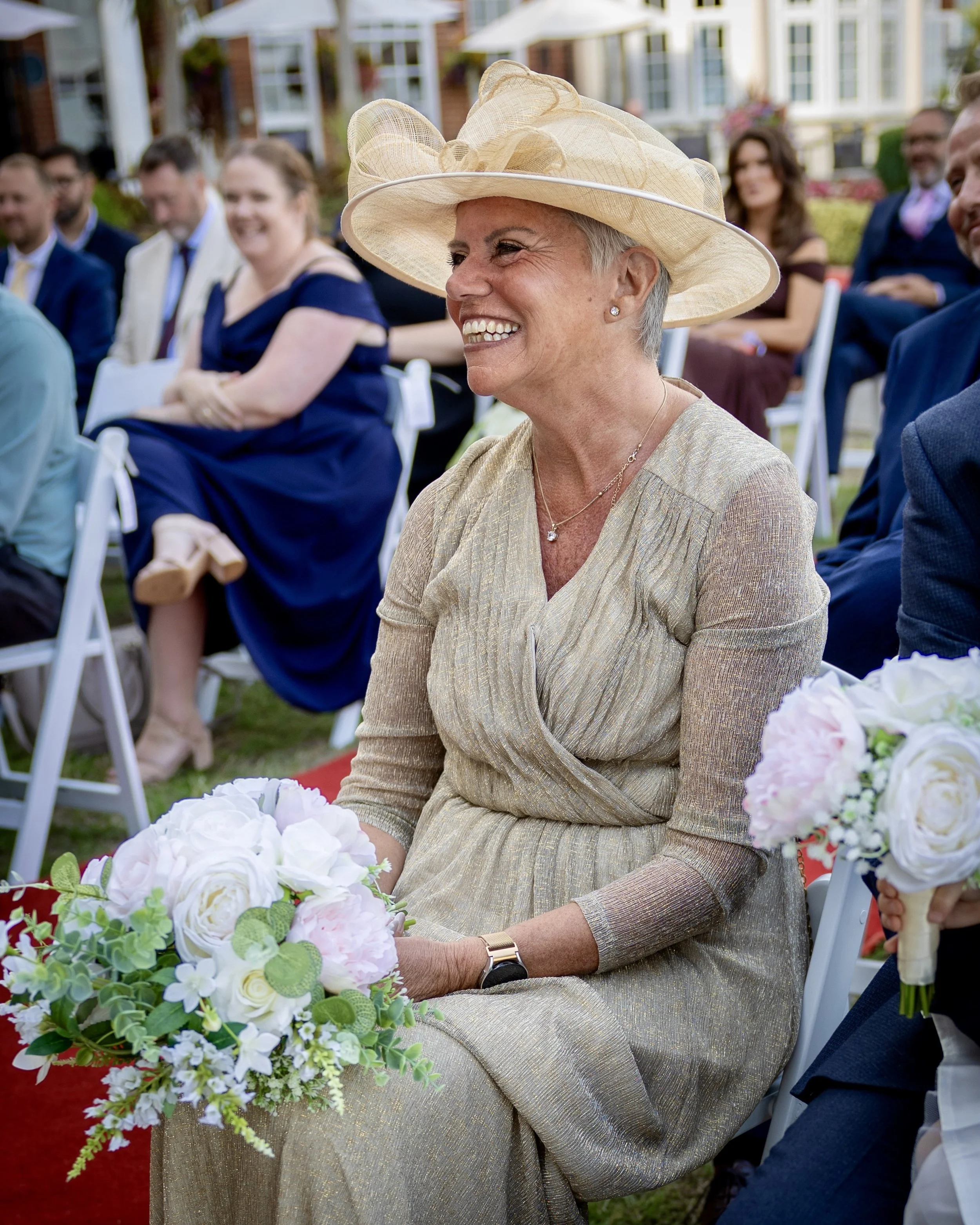 An elderly woman with short gray hair wearing a large beige hat, a gold dress, and jewelry, smiling while sitting at a wedding or formal outdoor event, with a bouquet of white and green flowers in front of her, surrounded by seated guests outdoors.