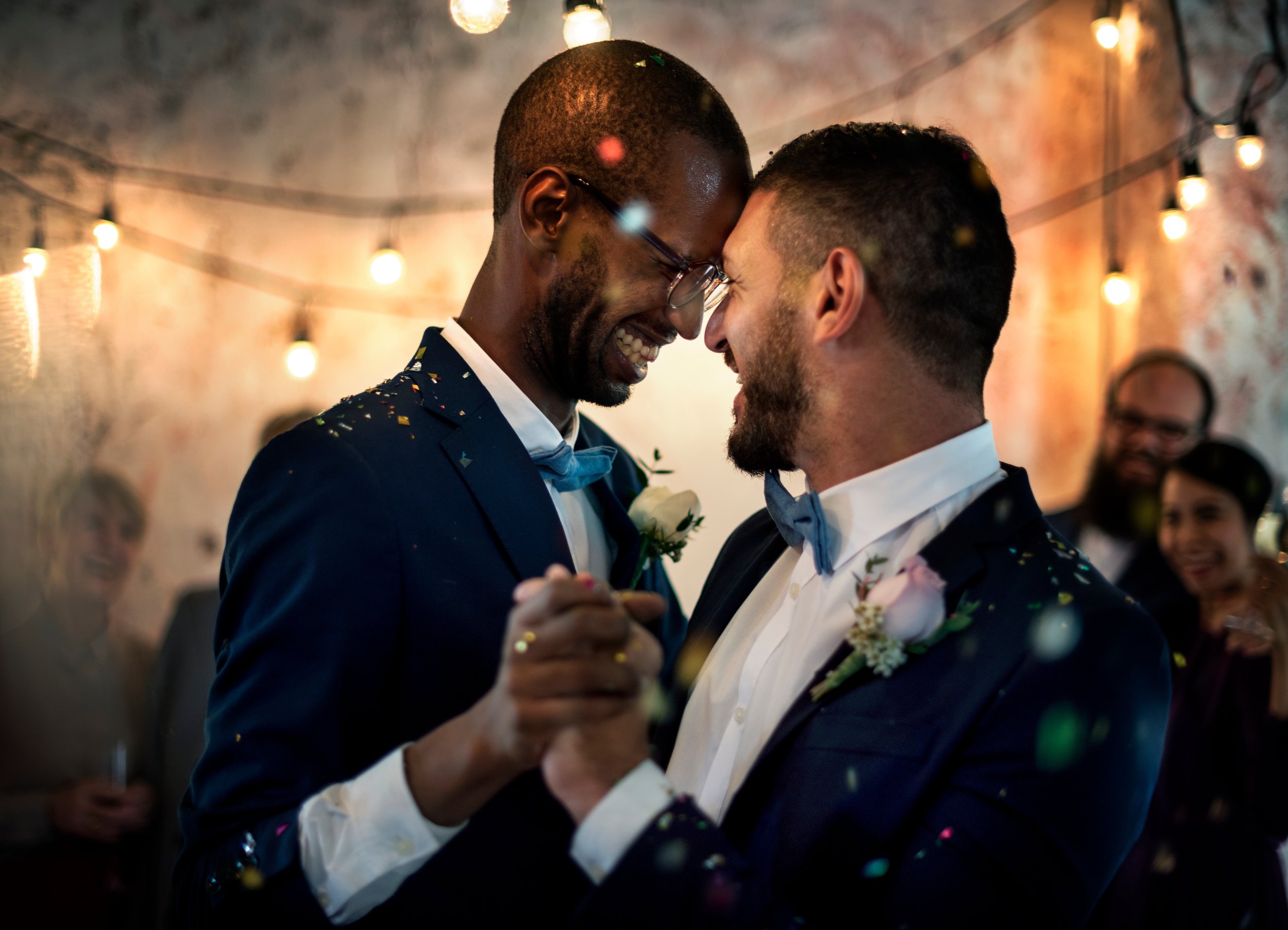 Two men in suits, holding hands, smiling and touching foreheads at a celebration with string lights and guests in the background.