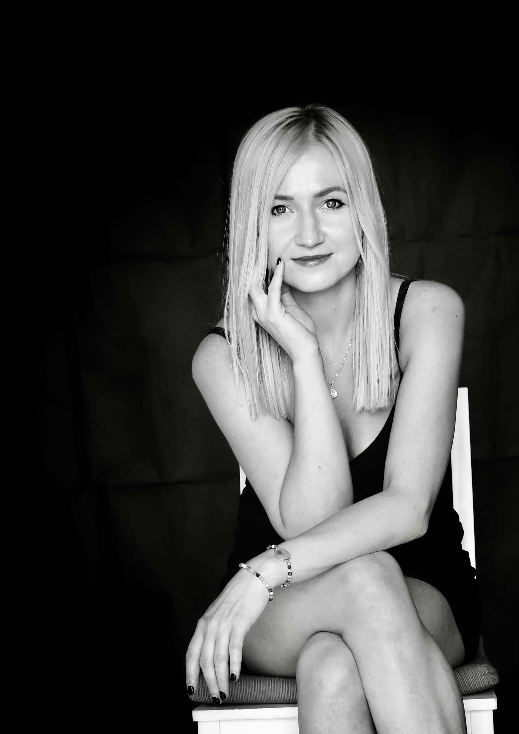 Black and white photo of a woman with long blonde hair, seated on a chair, wearing a sleeveless top, with one hand resting on her face and the other on her knee, against a dark background.
