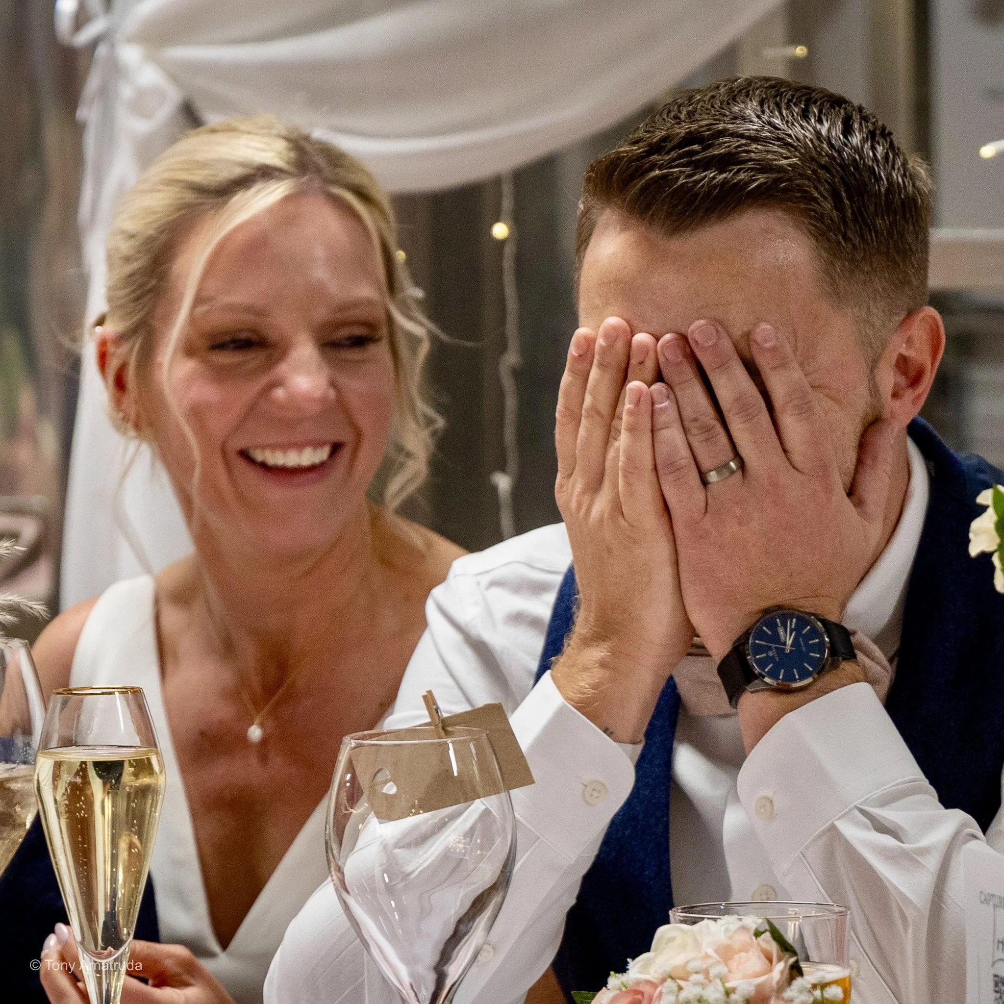 A woman and a man at a celebration, with the woman smiling and the man covering his face with his hands. There are glasses of champagne on the table in front of them.