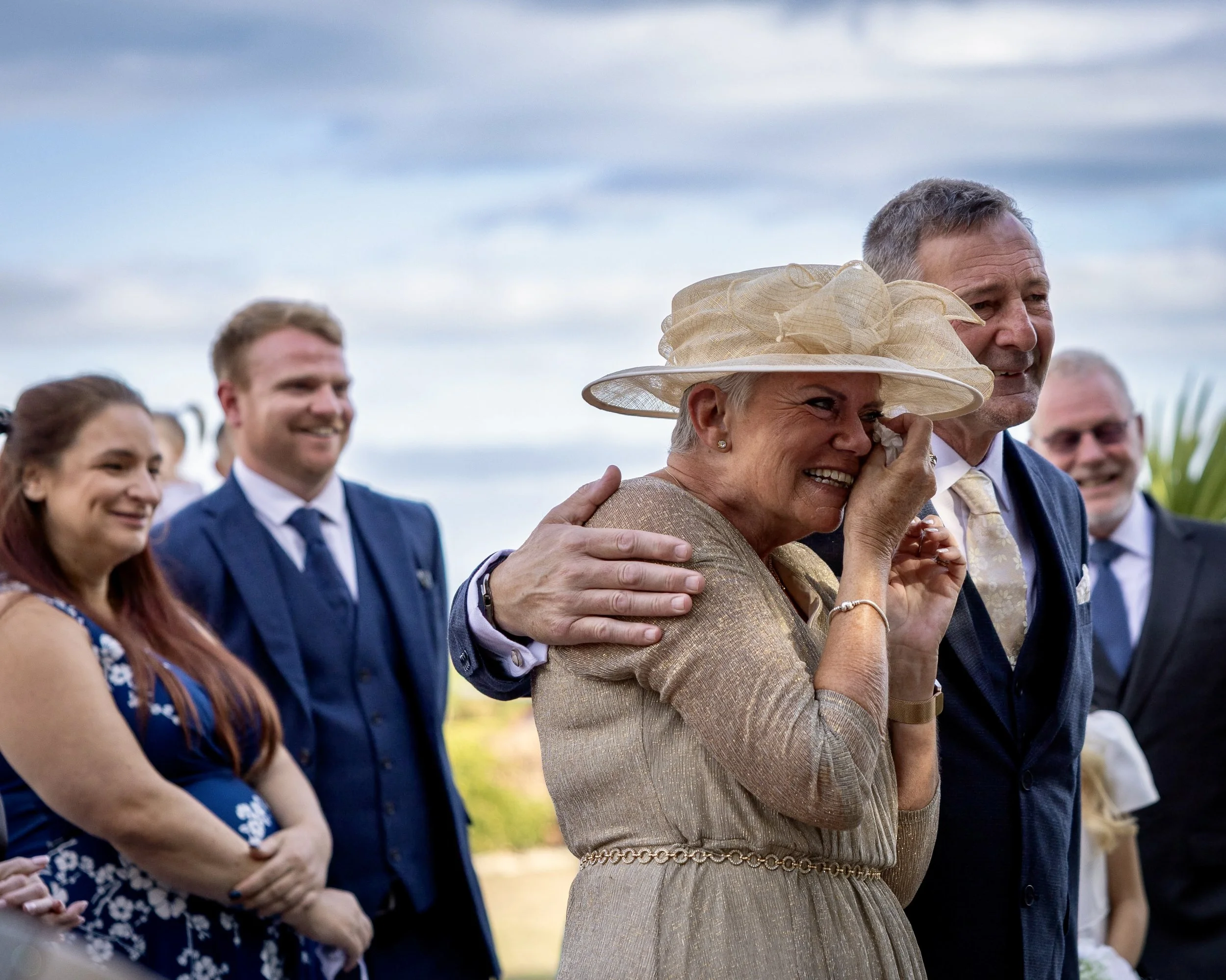 People at an outdoor event, with a woman wearing a large beige hat and a gold dress, talking and smiling with a man in a blue suit.