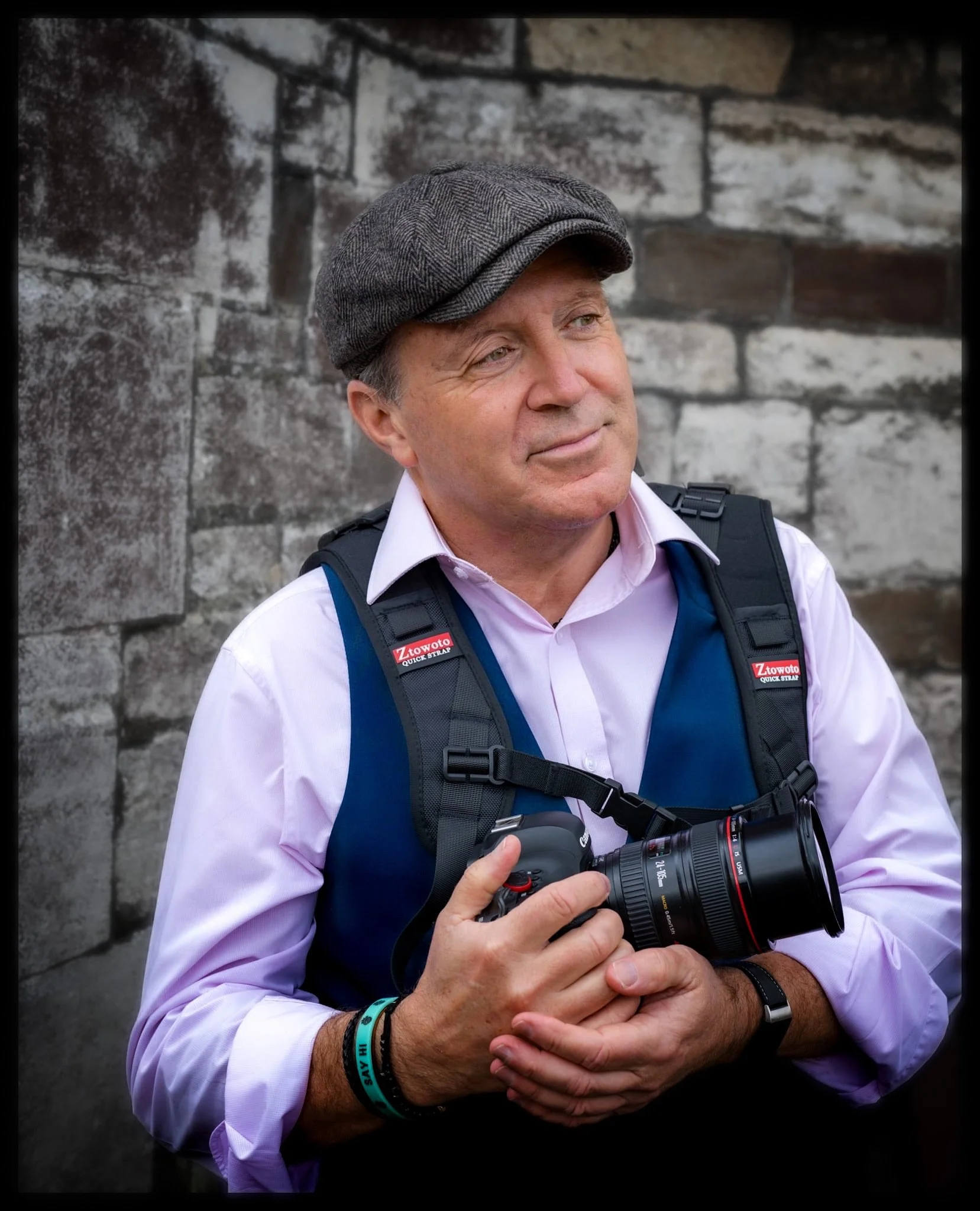 A man with a gray newsboy cap, wearing a light purple dress shirt, holding a camera, and wearing a black backpack. He is standing against a stone wall.