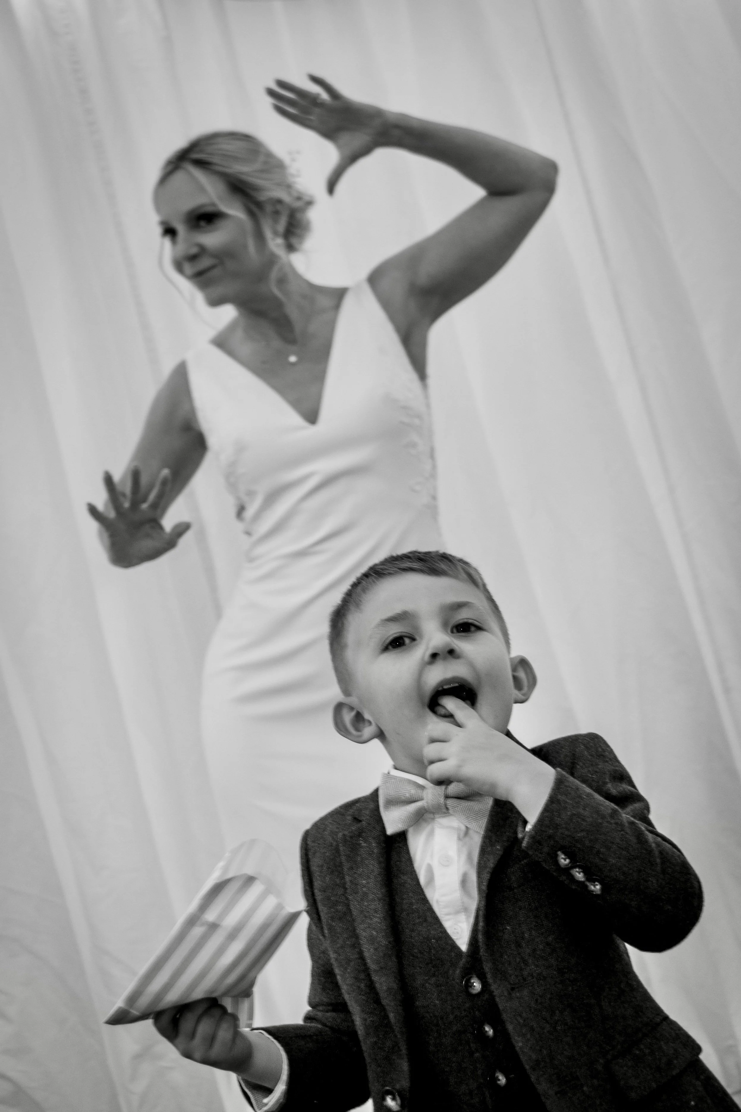 A woman with short blonde hair waving, holding a striped umbrella, and a young boy in a blazer holding his thumb to his mouth, standing in front of a plain wall.