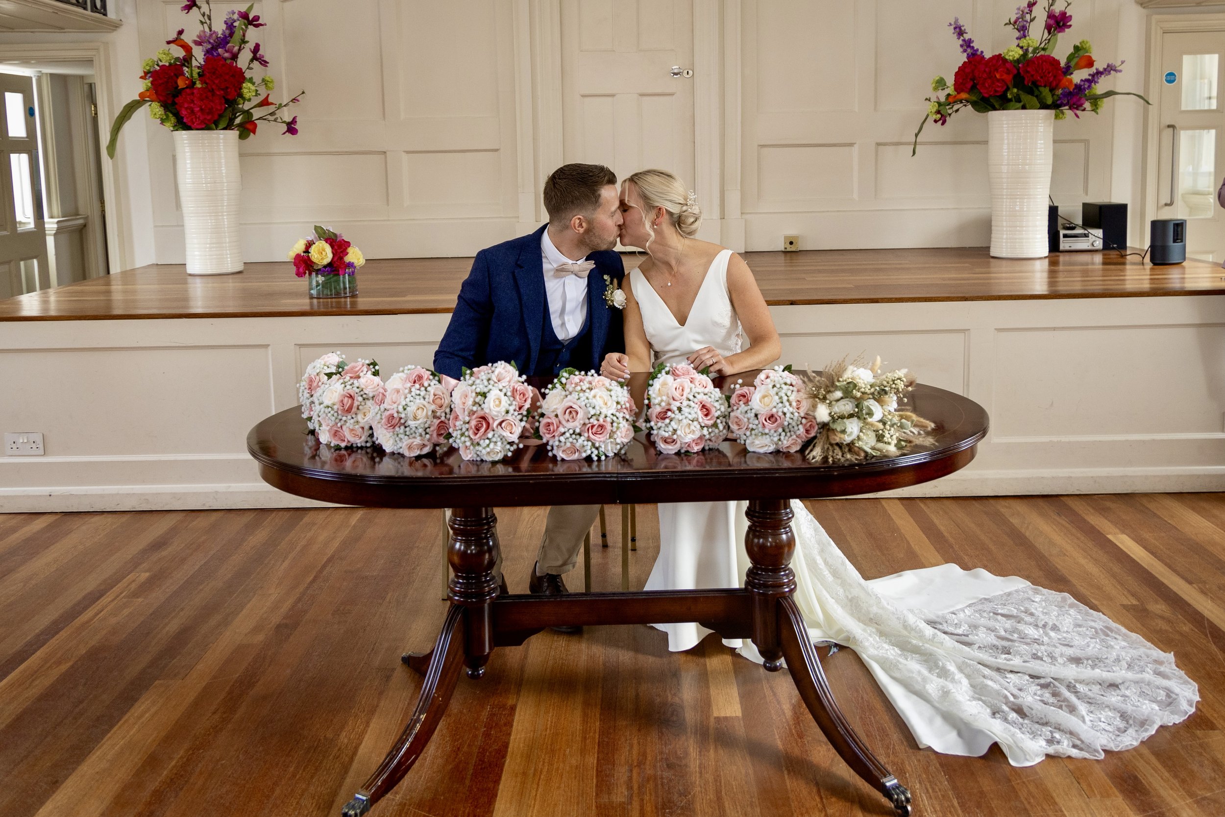 A bride and groom kissing at a wedding reception, seated in front of a table with pink and white floral arrangements, against a background of large floral vases on a stage.