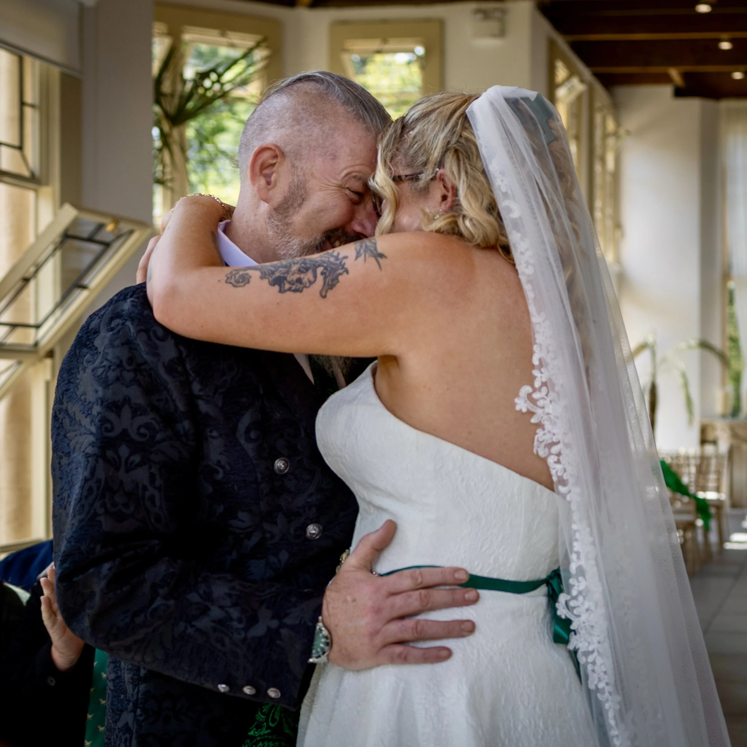 A couple hugging, possibly during a wedding or special event, with their cheeks pressed together, eyes closed, and arms wrapped around each other.