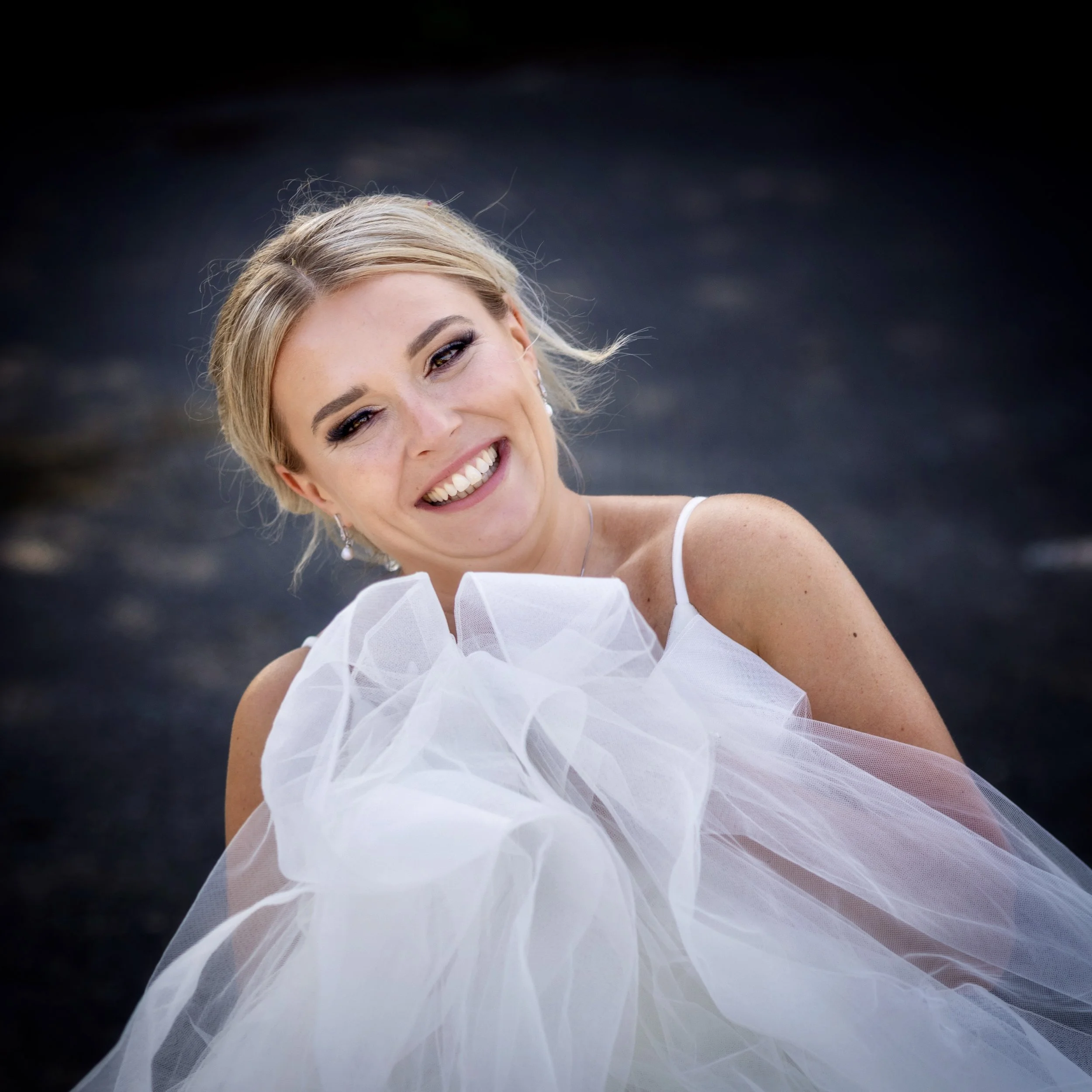 A smiling woman in a wedding dress holding a bouquet of white tulle, with a dark background.