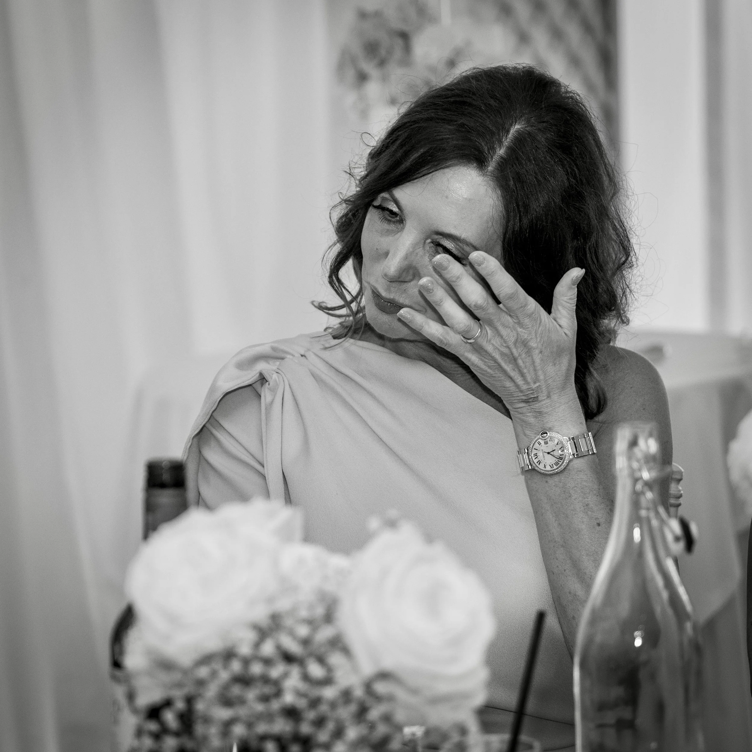 A woman with dark wavy hair, wearing a watch, touches her face with her hand, appearing emotional. She is at a table with flowers and a bottle in the foreground, in a light-colored setting.
