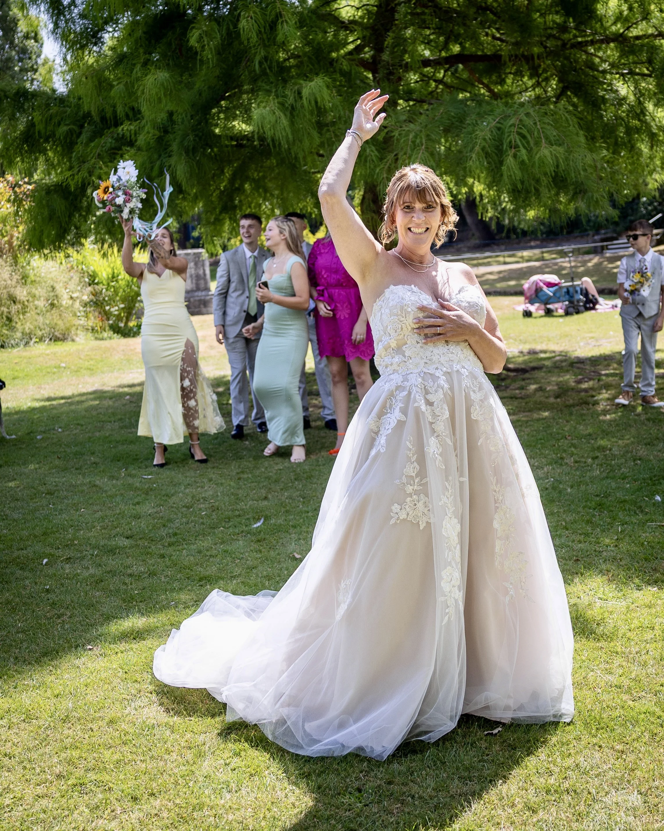 Smiling bride in a white lace wedding dress standing on grass in a park, with friends and family in the background during a wedding celebration.