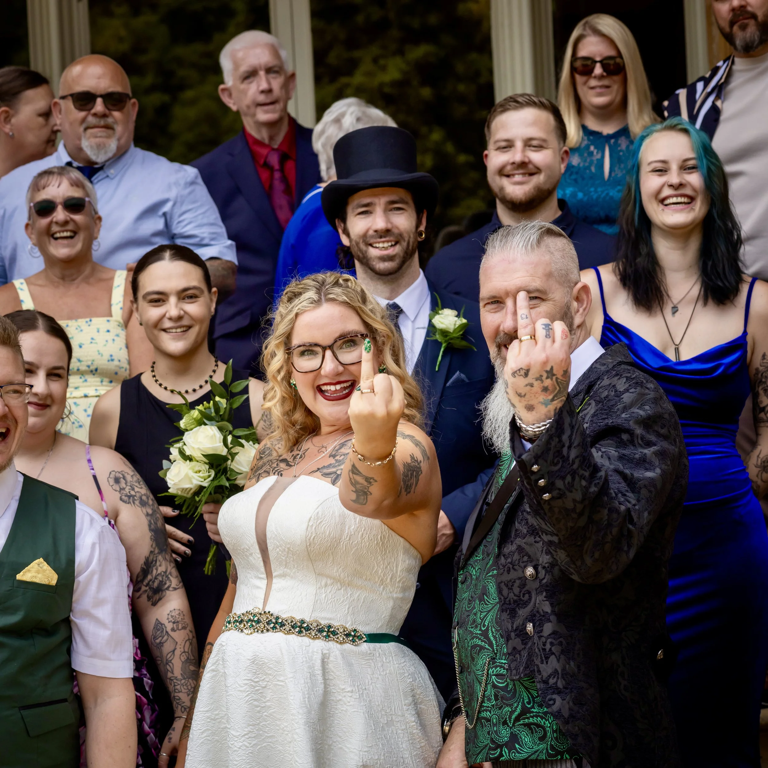 A group of smiling diverse people gathered for a celebration outdoors, with some wearing formal attire and others in casual clothes, including a woman with blue hair and a man with tattoos.