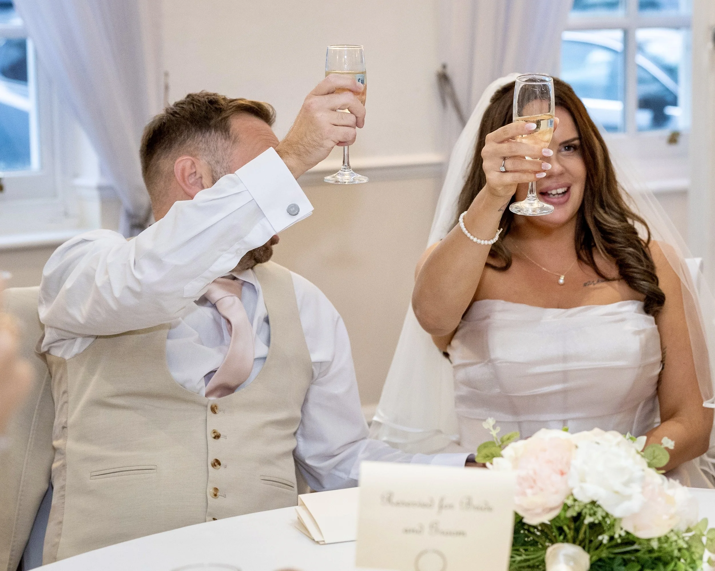 A newlywed couple dressed in wedding attire raising glasses of champagne for a toast at their wedding reception.
