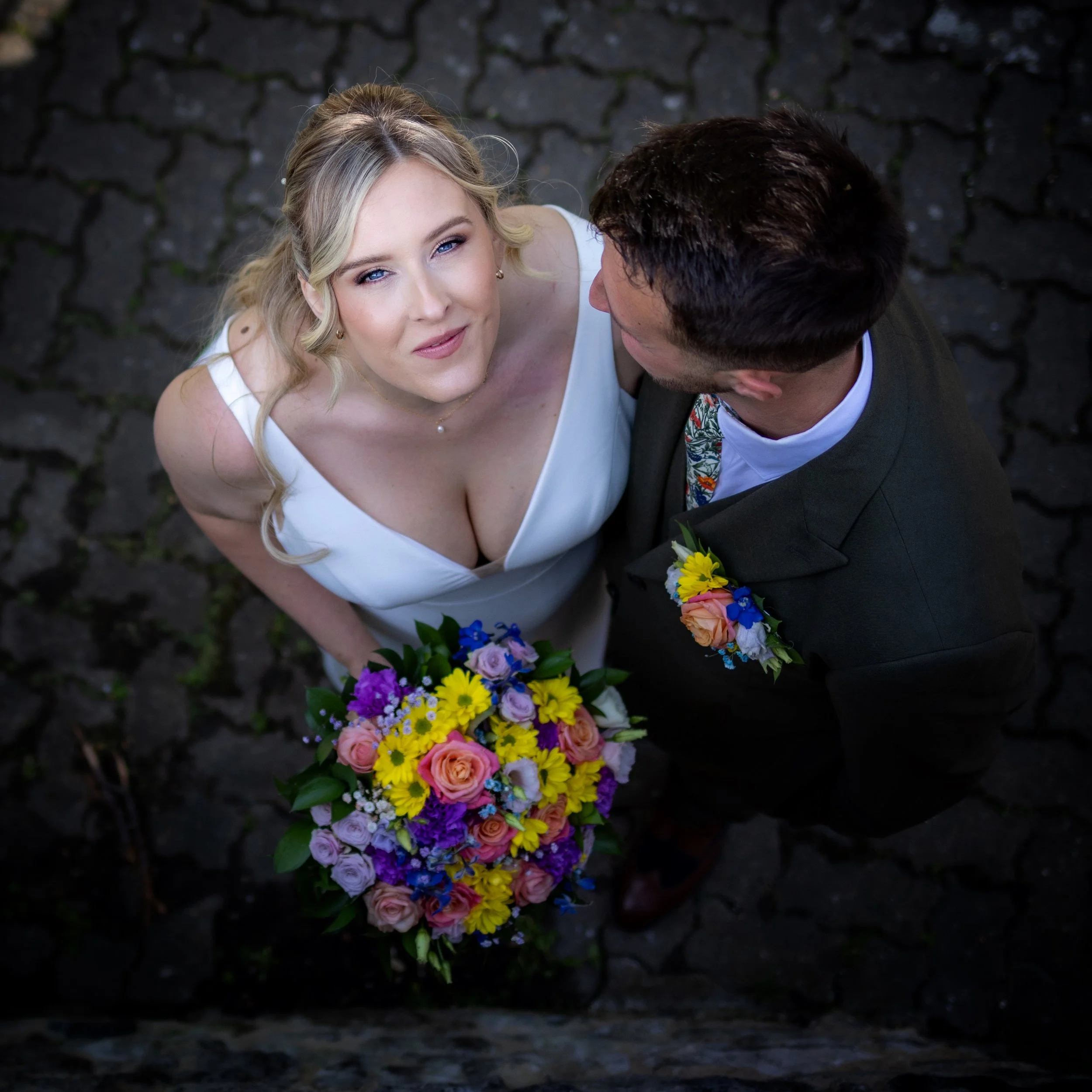 A bride with blonde hair in a white dress holding a colorful bouquet looks up at the camera, standing beside a groom in a black suit with a boutonniere, on a dark cobblestone path.