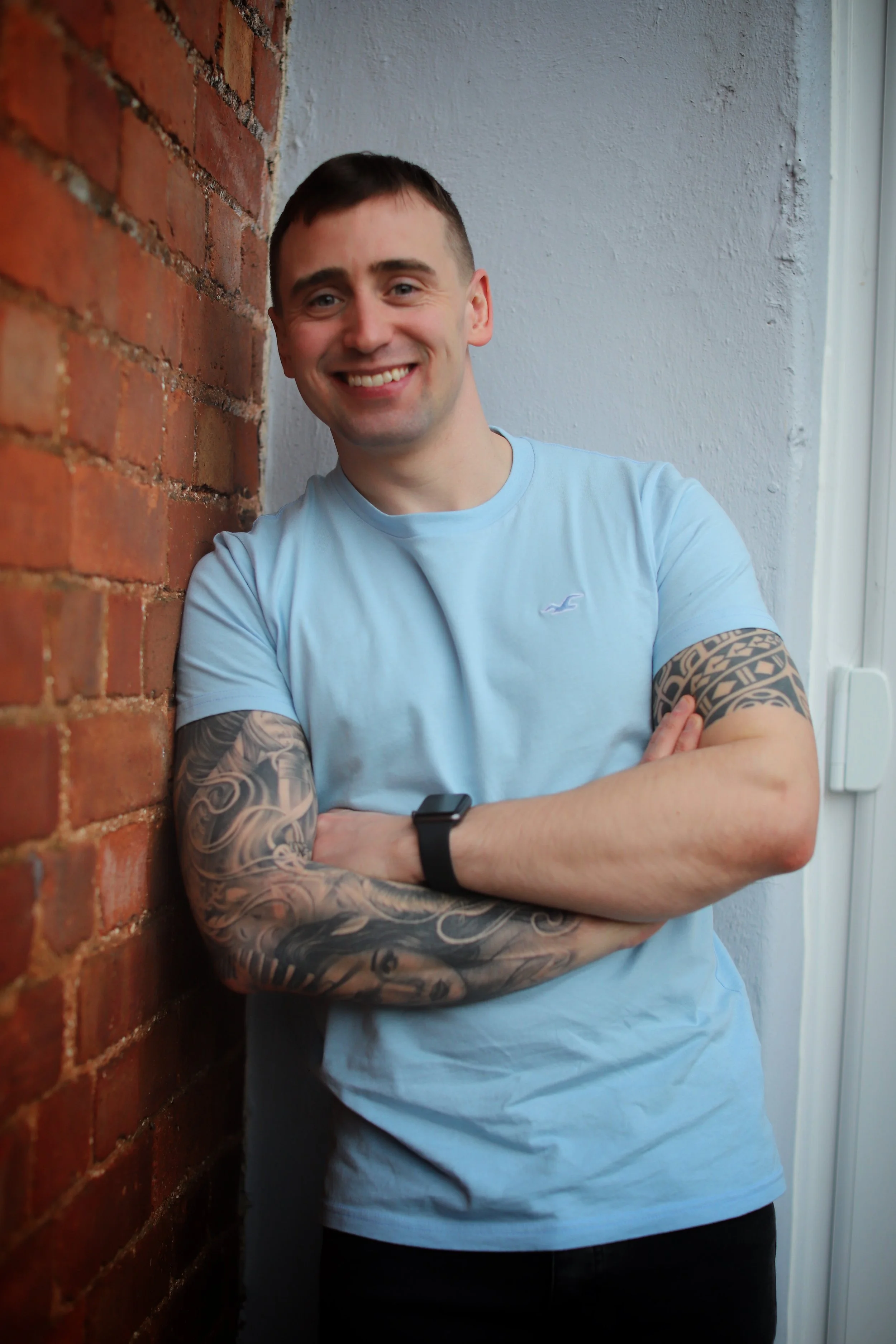 Smiling man with tattoos on arms, wearing a light blue T-shirt, leaning against a brick wall and a white wall.