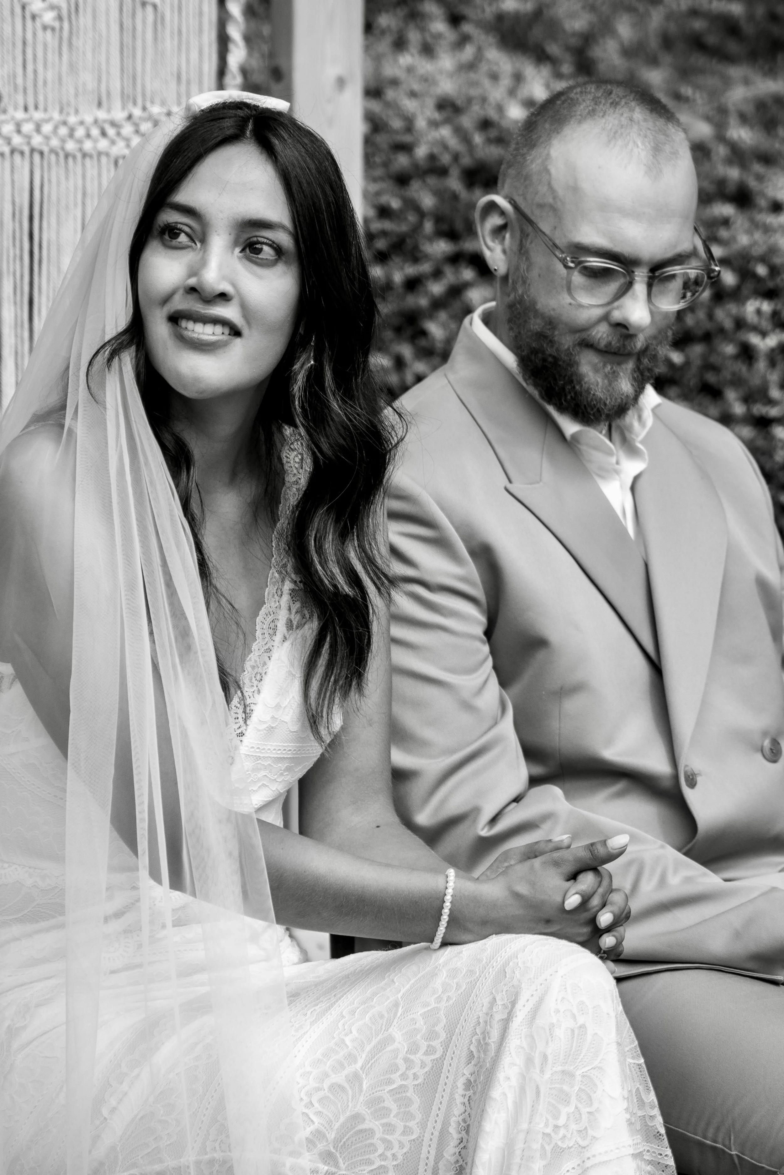 A black and white photo of a bride and groom sitting outdoors, holding hands, with the bride looking to the side and smiling gently, and the groom looking down with glasses and a beard.