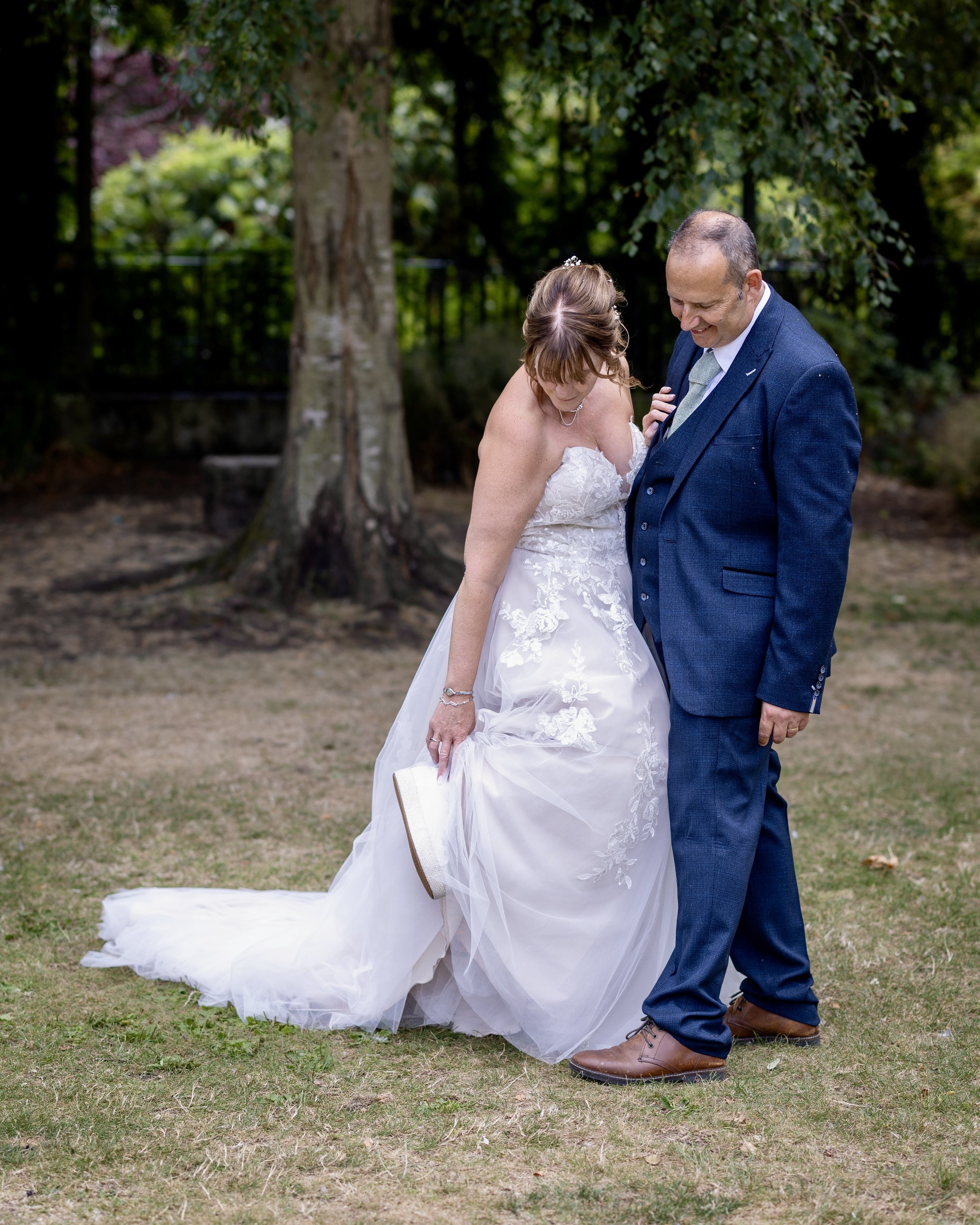 A bride and groom dancing outdoors on grass with trees in the background. The bride is in a white wedding gown and lifting her leg, showing beige shoes. The groom is in a dark blue suit and brown shoes, smiling as they dance.